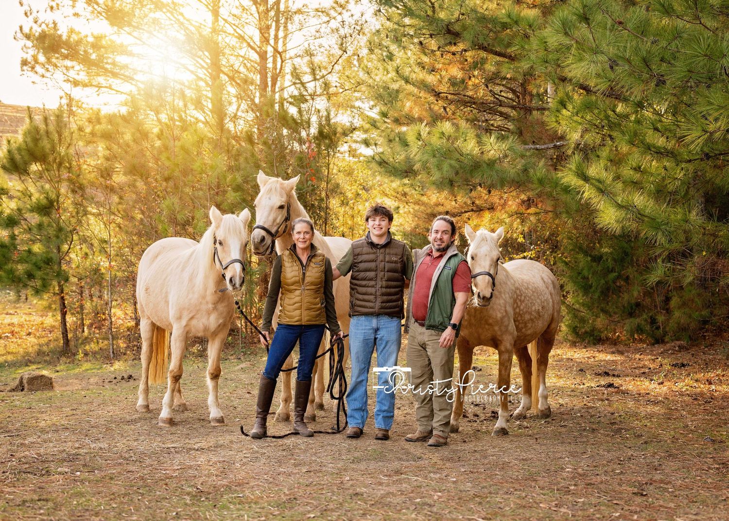 Two people and a man stand with three palomino horses in a sunny forest clearing during golden hour.