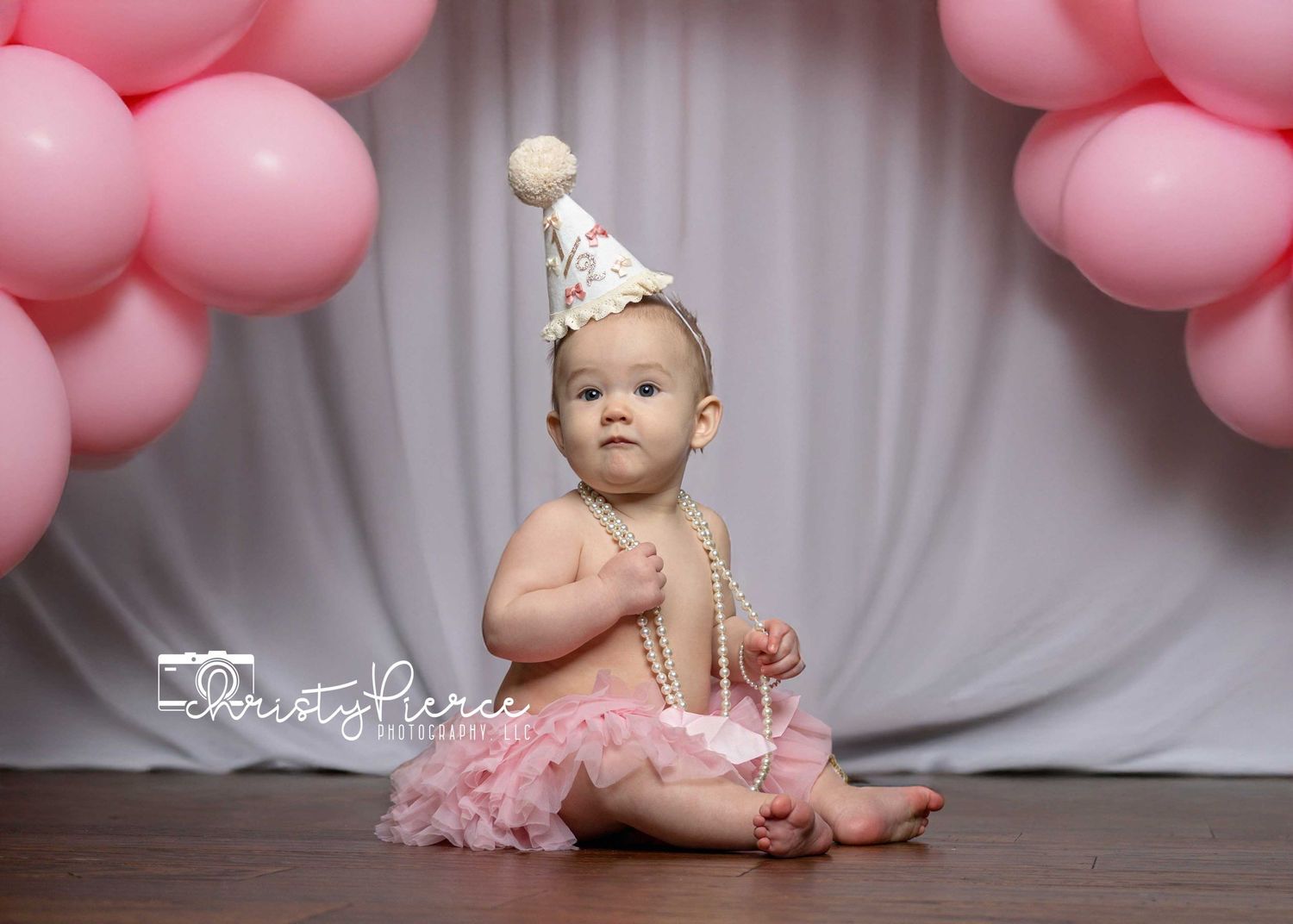 Baby in pink tutu and party hat sits on wooden floor with pink balloon arch decorations in background.