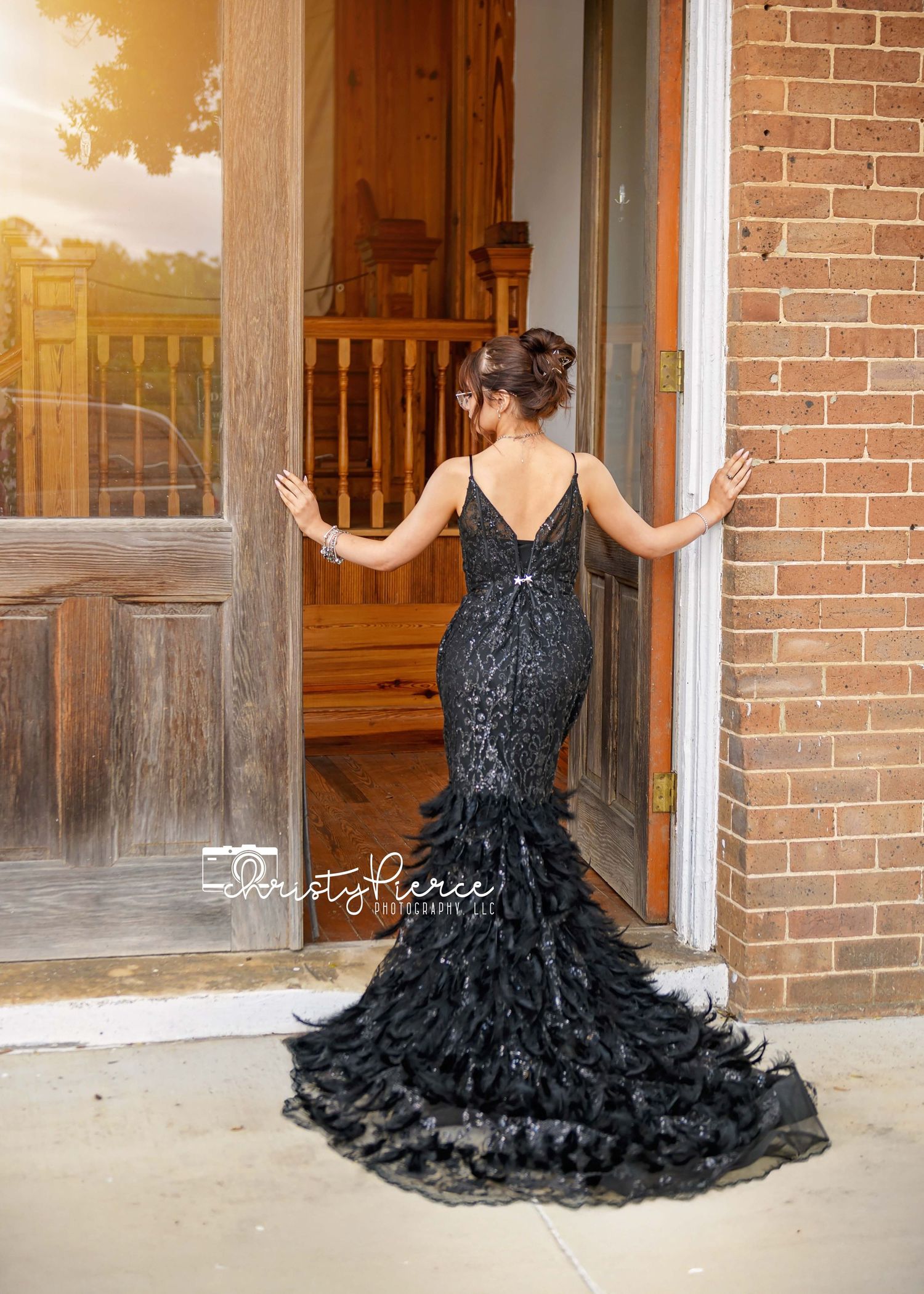 A woman in a black feathered evening gown poses elegantly in front of a brick building at sunset.