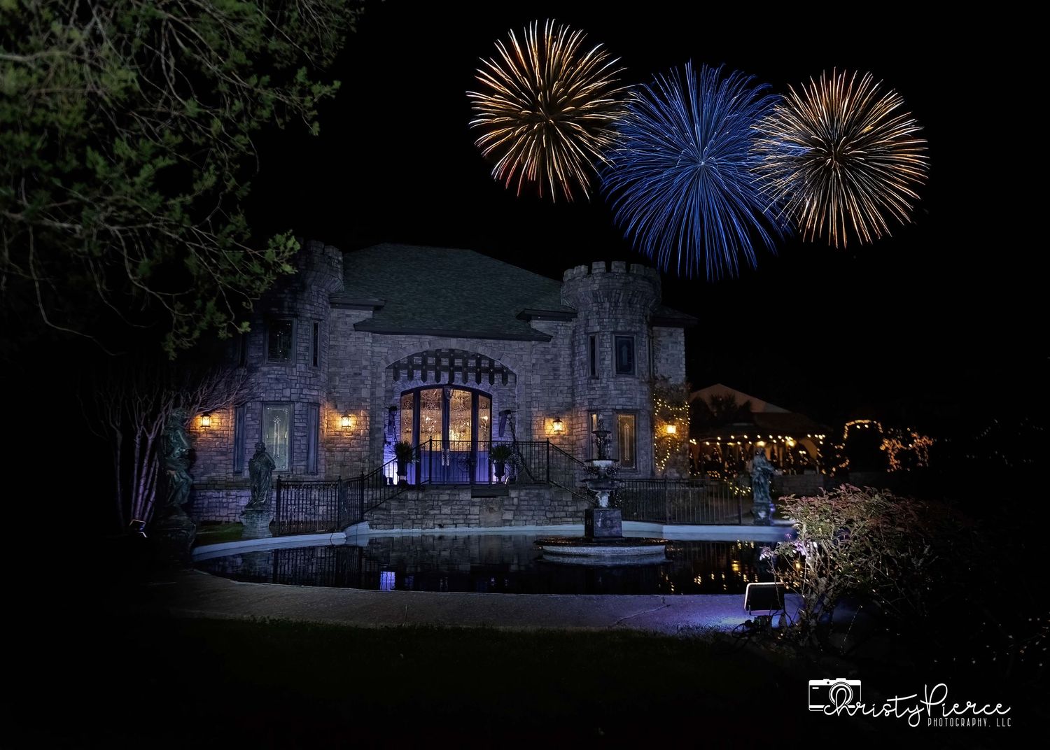 Elegant stone mansion illuminated at night with fireworks bursting in blue and gold above the building.