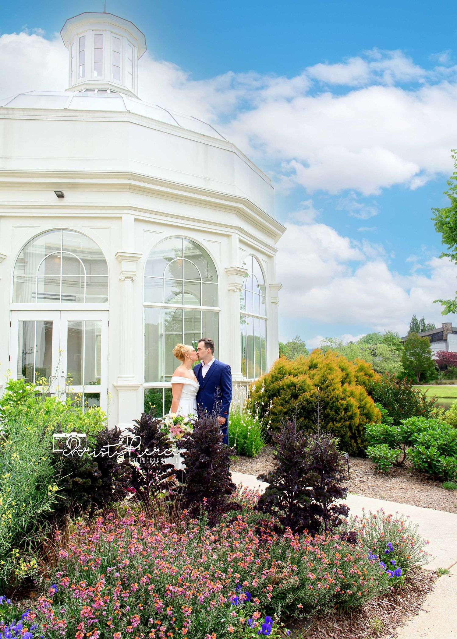 A wedding couple shares a romantic kiss in a lush garden beside an elegant white glass conservatory under a bright blue sky.