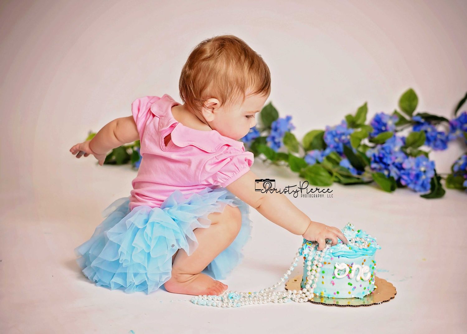 Baby in pink top and blue tutu reaching for small decorated cake with blue flowers in background.