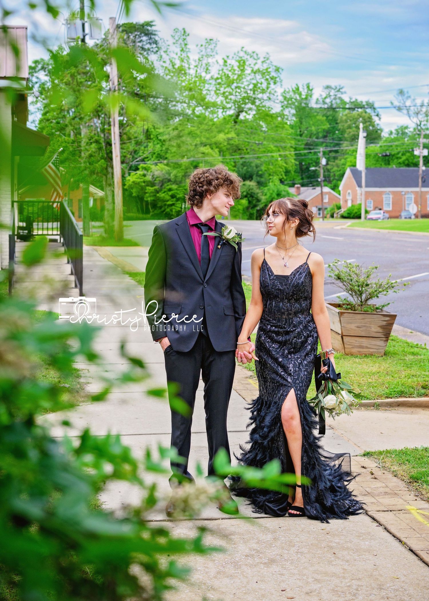 A couple in formal attire poses together outdoors on a spring day with blooming trees and greenery in the background.