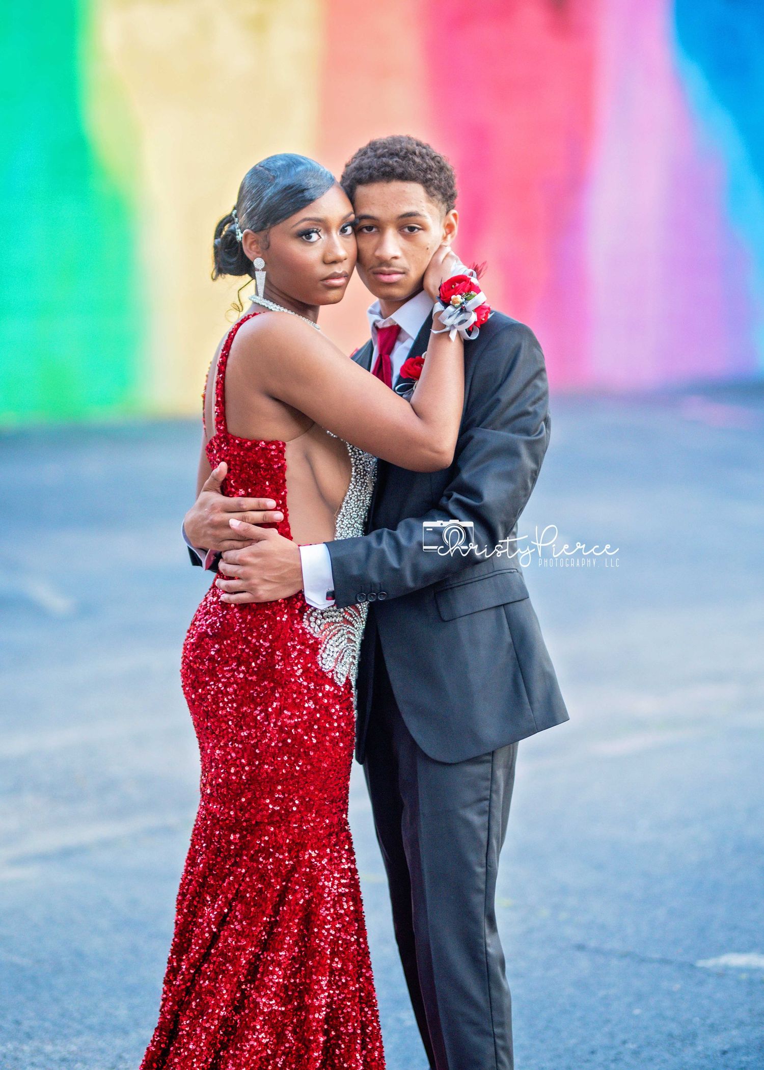 A couple in formal attire pose for prom photos, with the person in a sparkling red gown and another in a black suit.