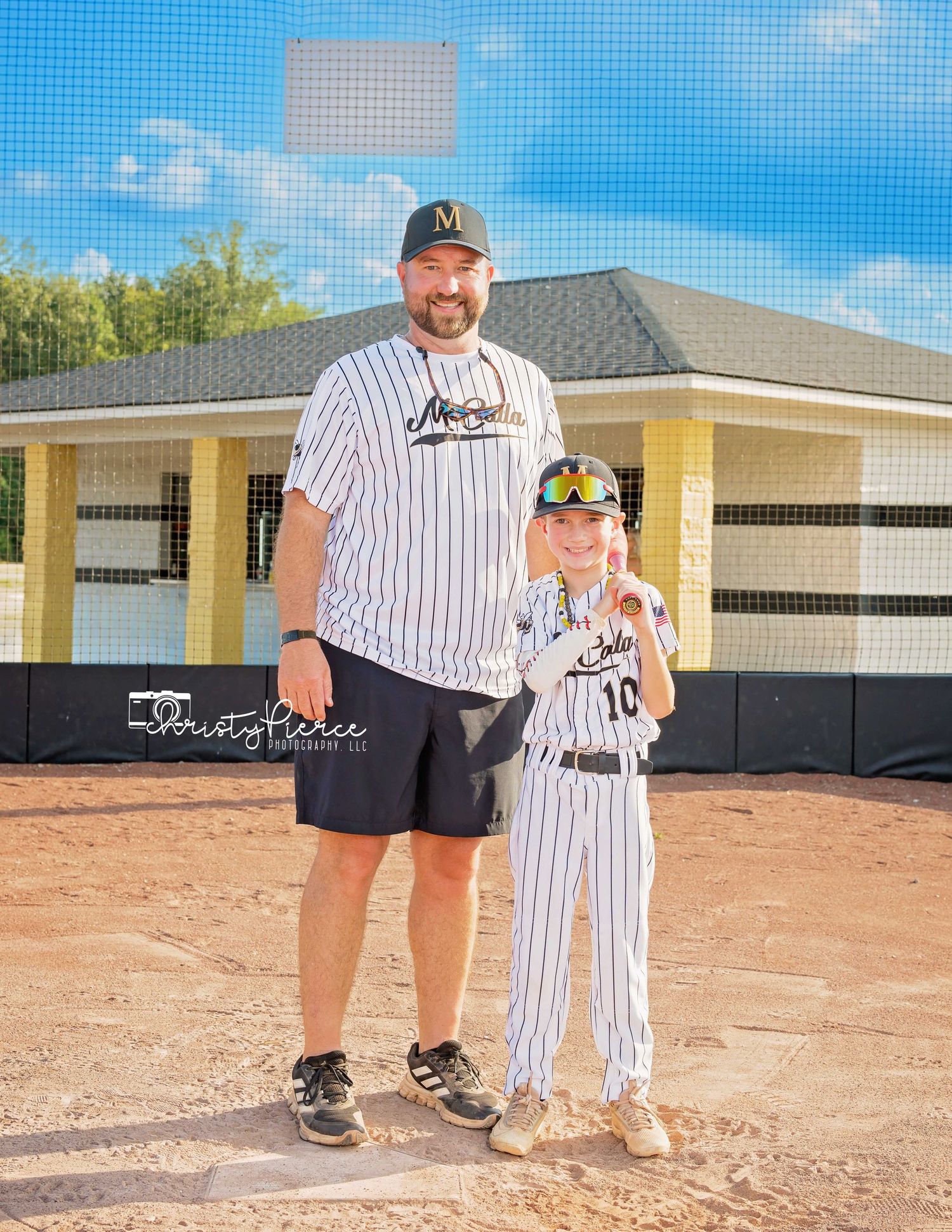 A baseball player and child stand together on a dirt field wearing matching pinstriped uniforms.