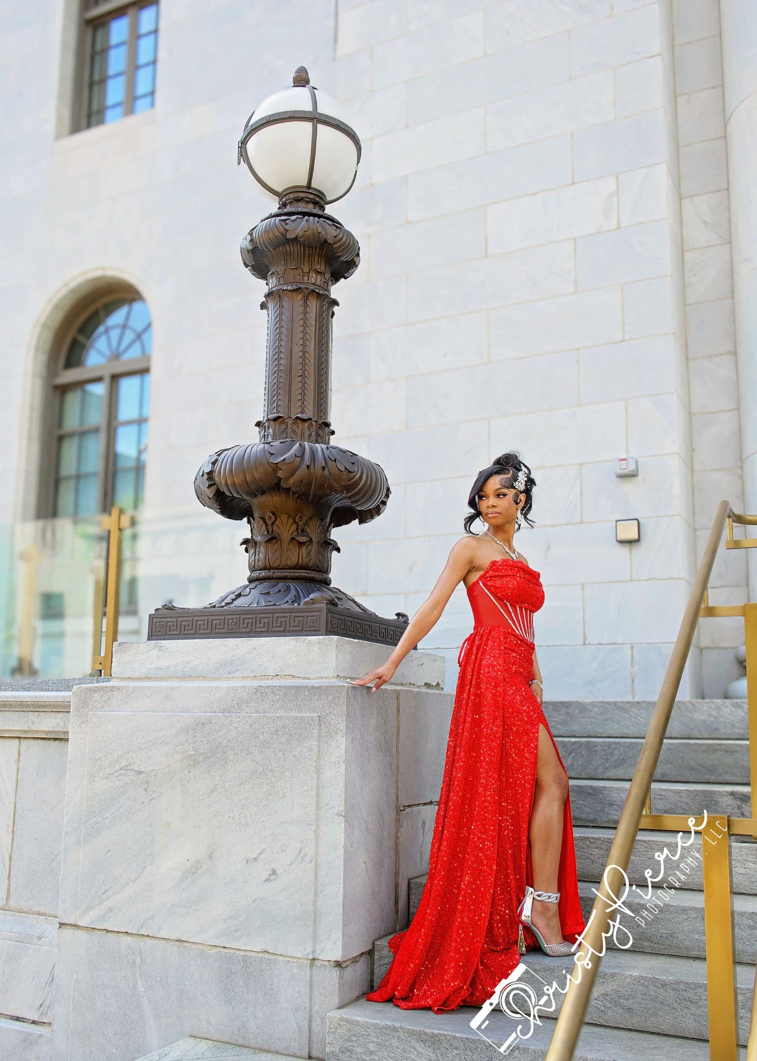 Woman in red gown poses elegantly on stone steps beside ornate lamp post at grand building entrance.