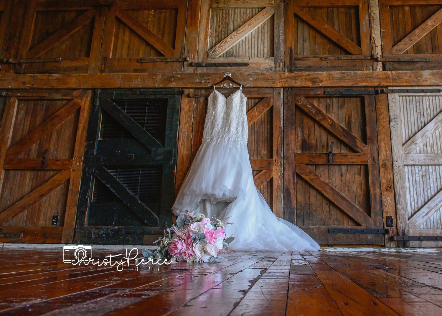 Elegant lace wedding dress hanging on rustic barn doors with a pink bridal bouquet on the wooden floor.