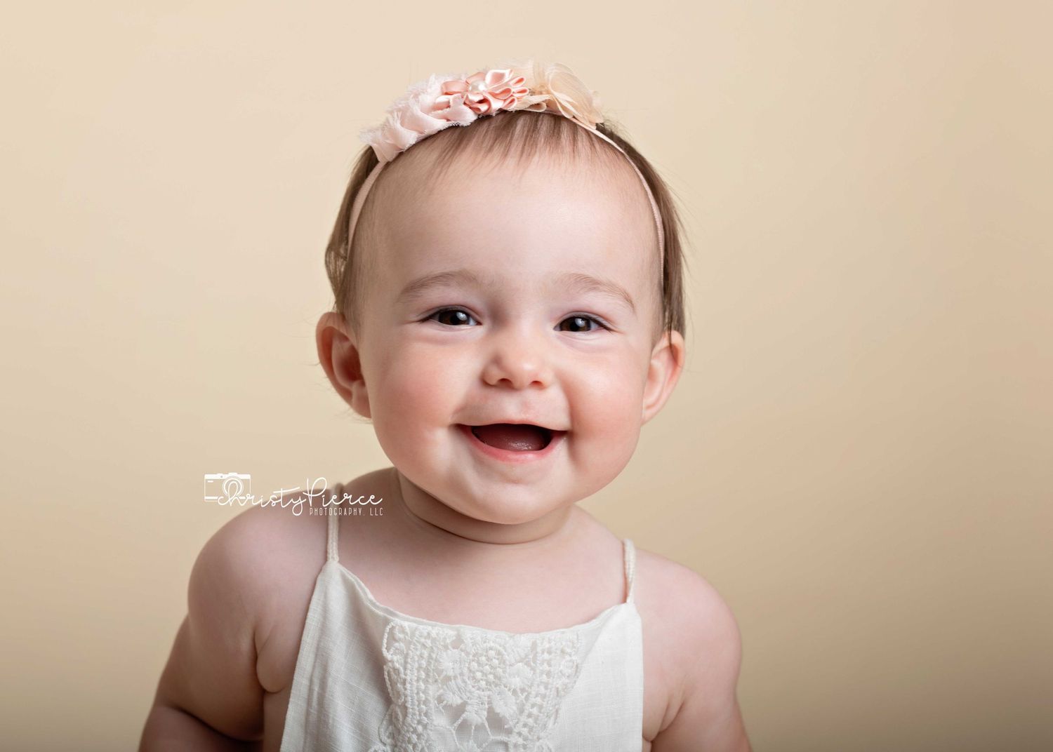 A smiling baby in a white lace dress and pink floral headband poses against a beige background.