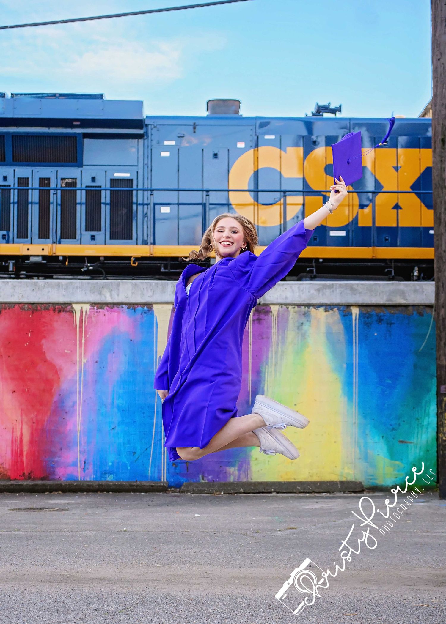 A jubilant graduate in purple cap and gown jumps joyfully in front of a colorful mural with a CSX train in the background.