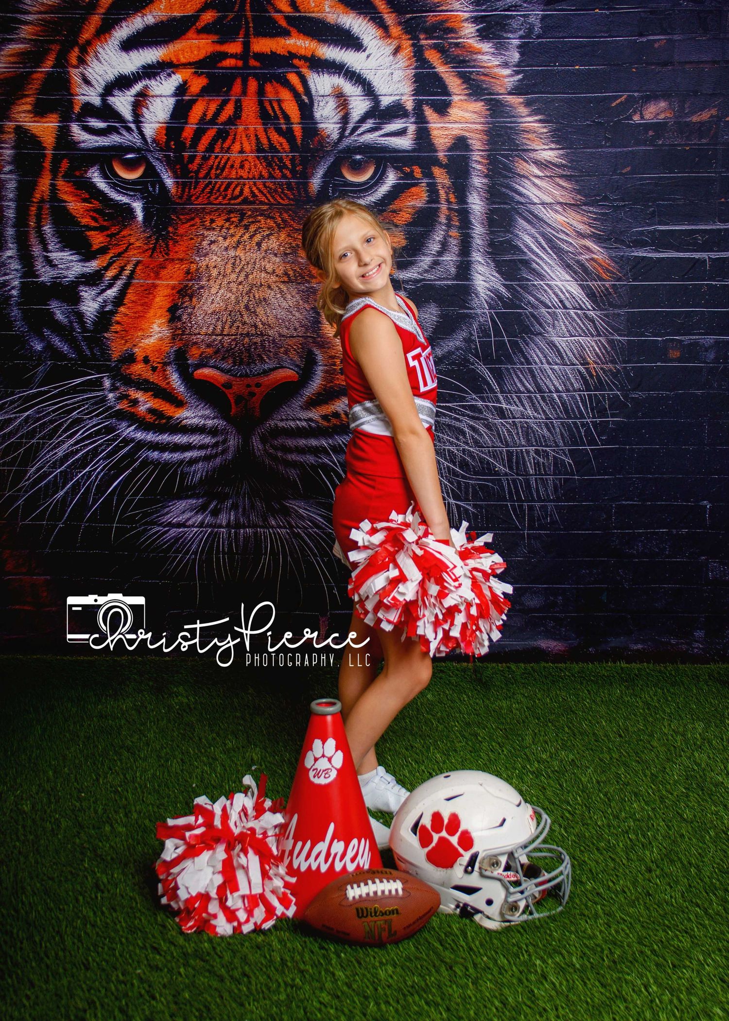 Young cheerleader in red uniform poses with pom-poms, football helmet, and megaphone in front of a tiger mural backdrop.