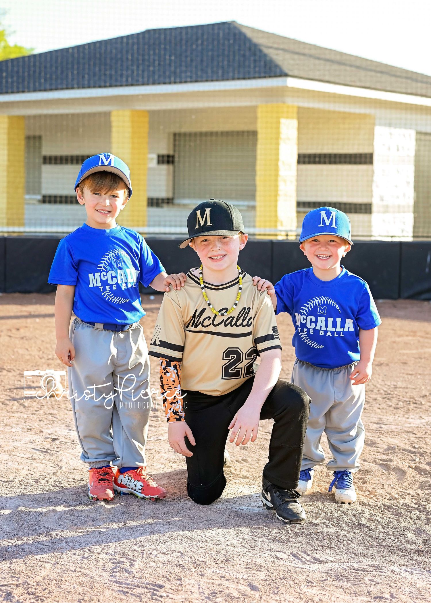 Three young baseball players in uniforms pose together on a dirt field with a yellow building in the background.
