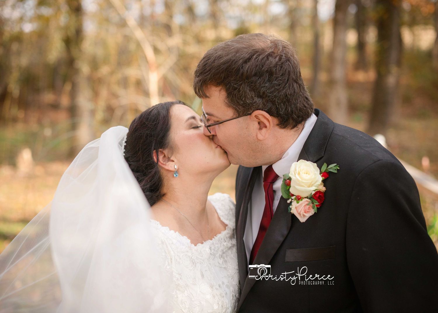 A bride and groom share a romantic kiss outdoors during their fall wedding ceremony with autumn foliage in the background.