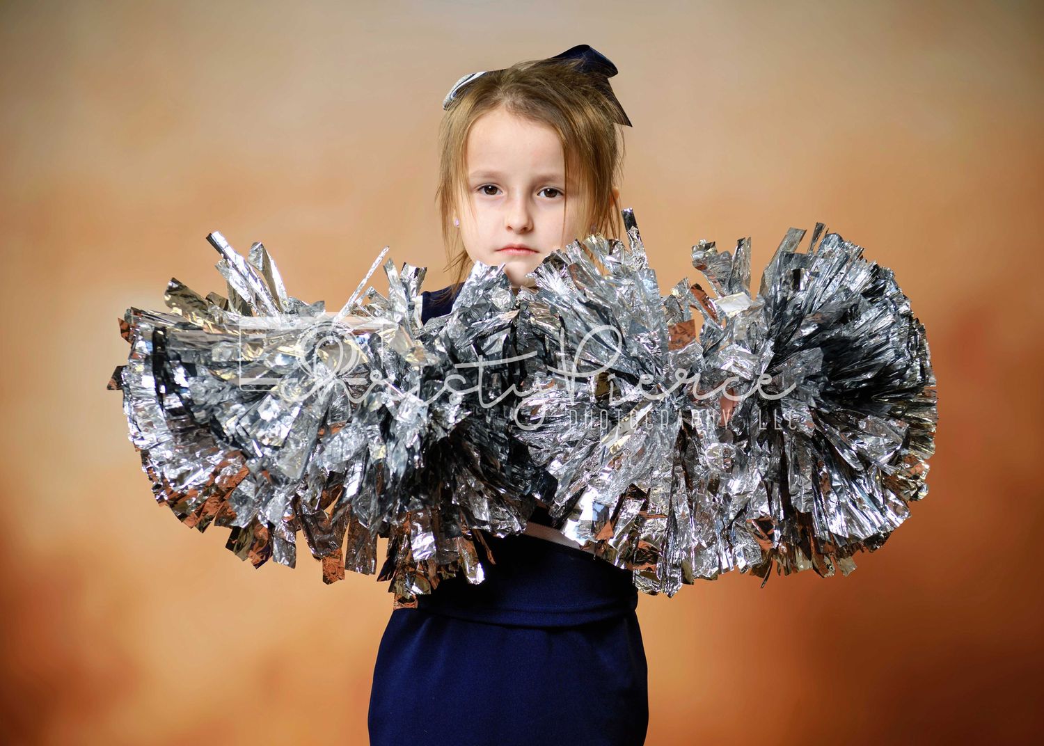 A cheerleader poses in a navy uniform while holding large silver pom poms against a brown backdrop.