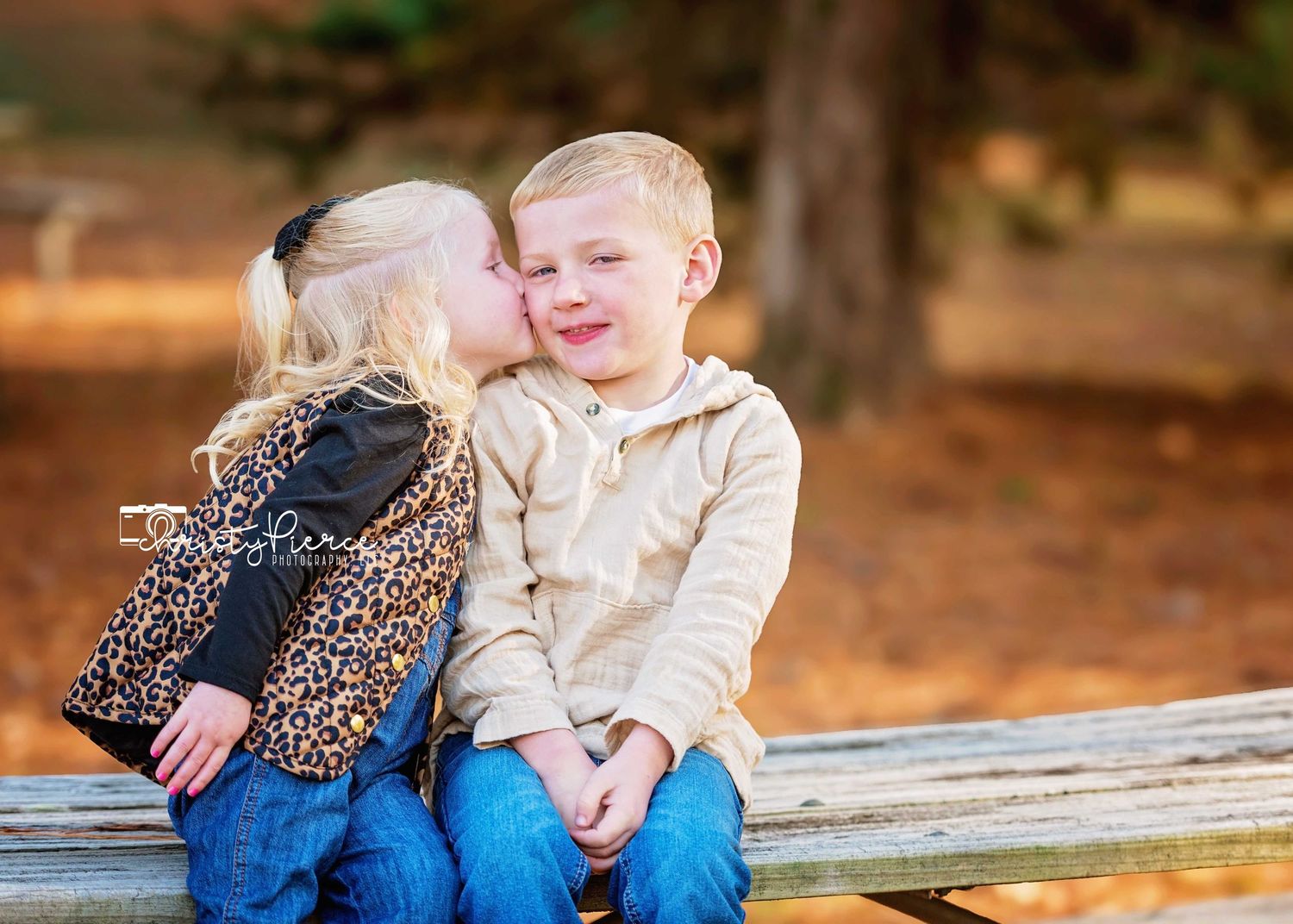Two young children share a sweet moment sitting together on a wooden bench outdoors in autumn.