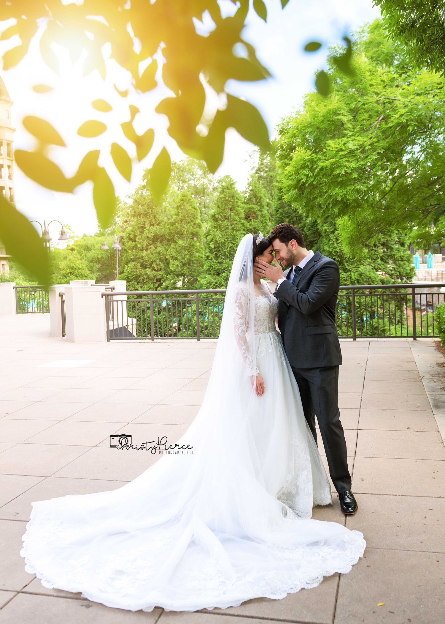 A romantic wedding couple embraces on a sunlit terrace surrounded by lush green trees and leaves.