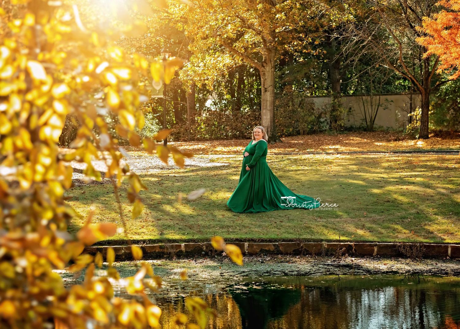 A person in an emerald green gown poses gracefully near a reflective pond surrounded by golden autumn foliage.