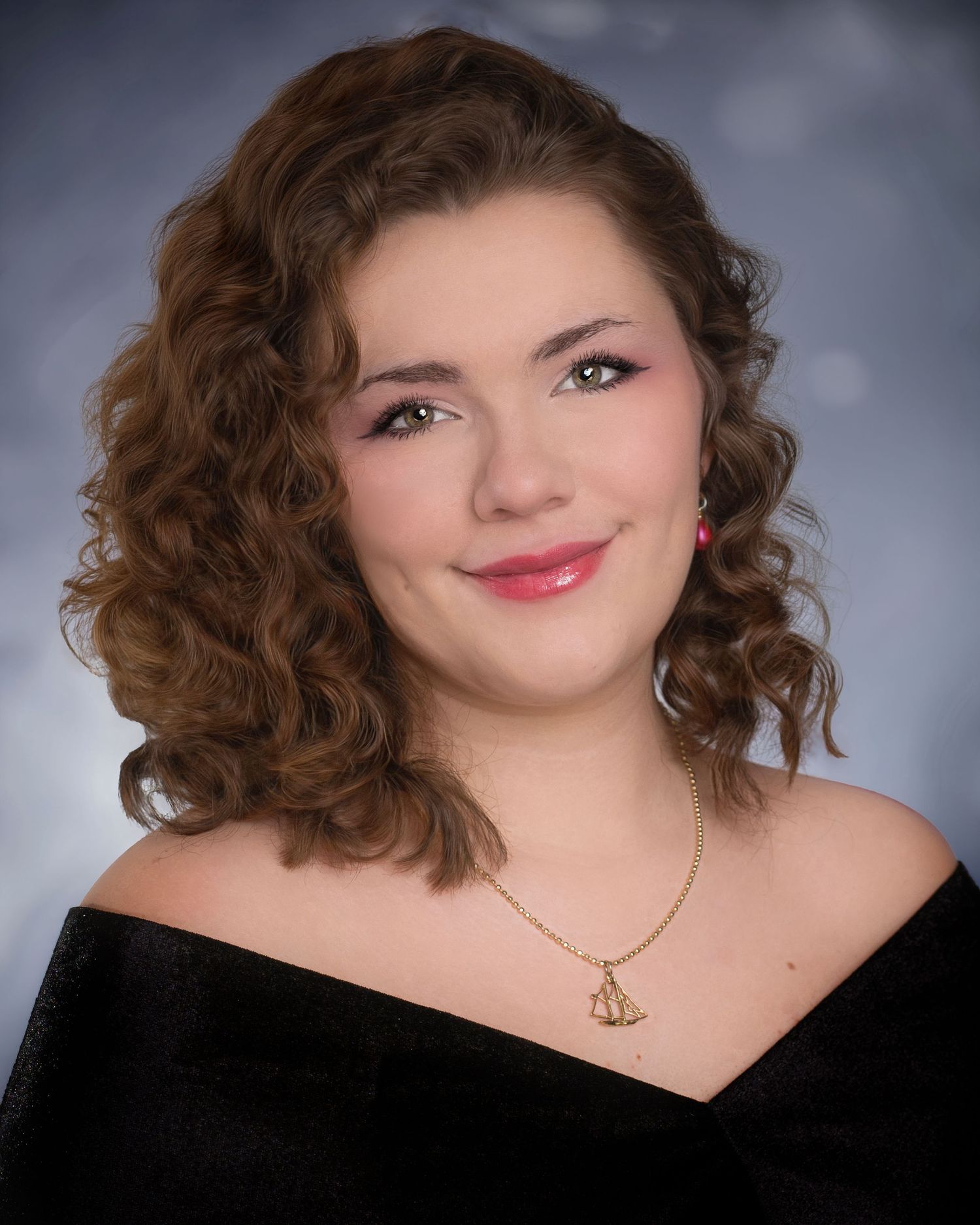 Young woman with curly auburn hair wearing a black off-shoulder top and gold sailboat necklace in a professional portrait.