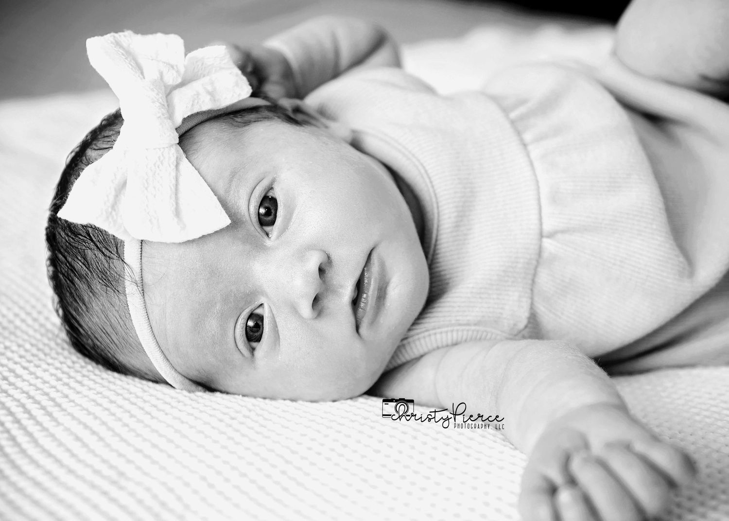 A black and white portrait of a baby wearing a white bow headband while lying on textured fabric.
