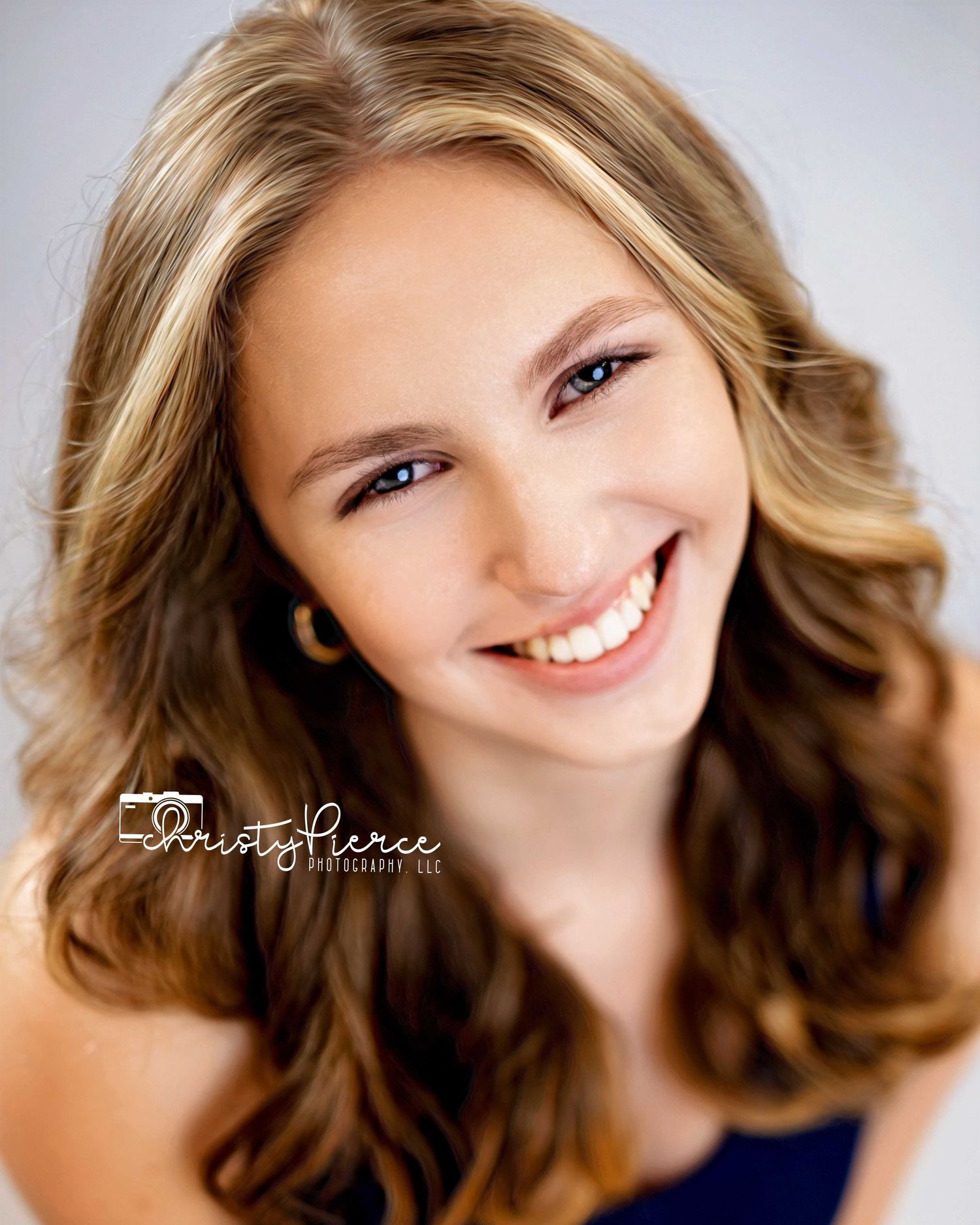 Professional portrait photo showing a bright smile and wavy brown hair against a soft gray background.
