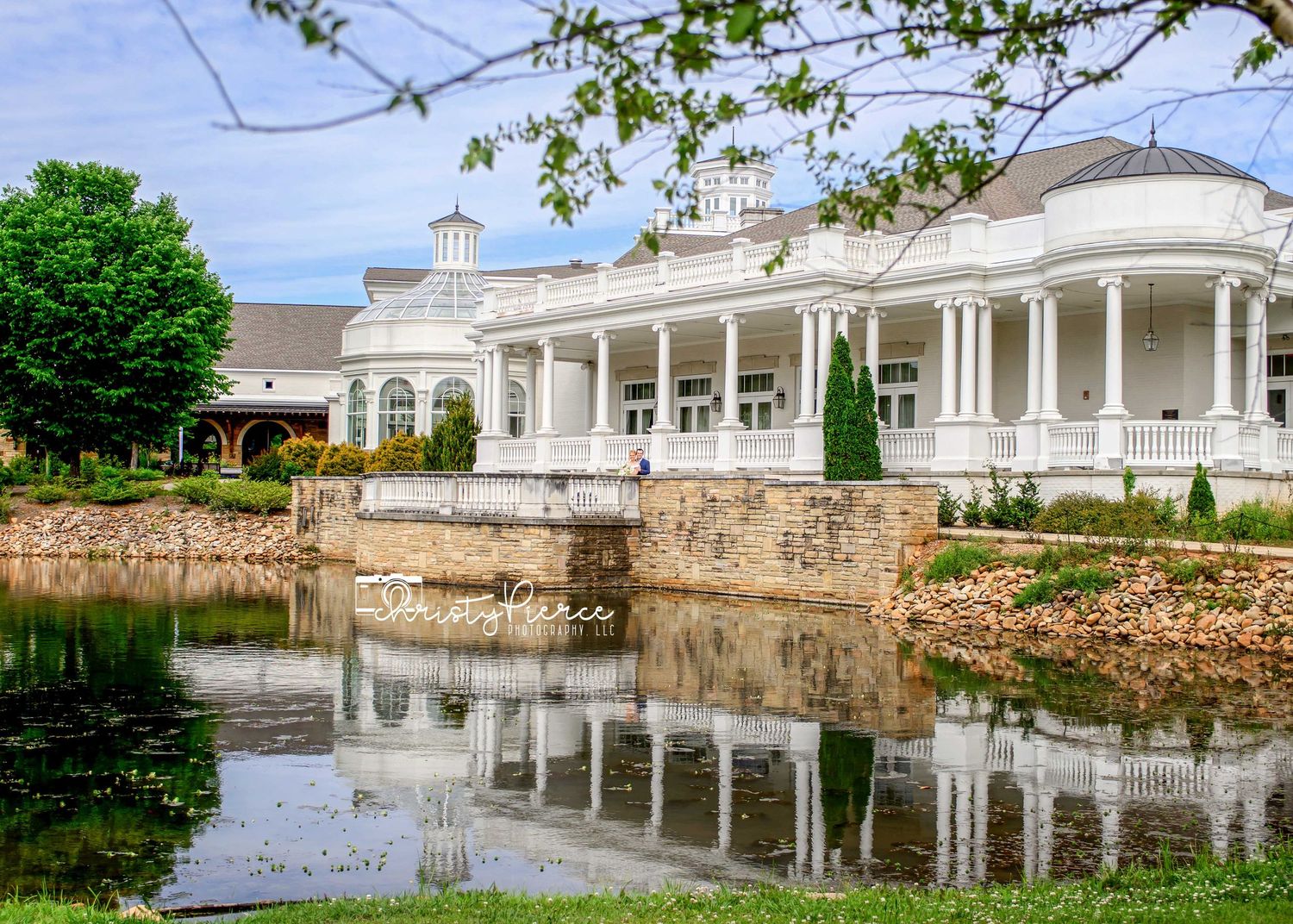 Elegant white colonial mansion with grand columns reflecting in a serene pond, surrounded by lush greenery.