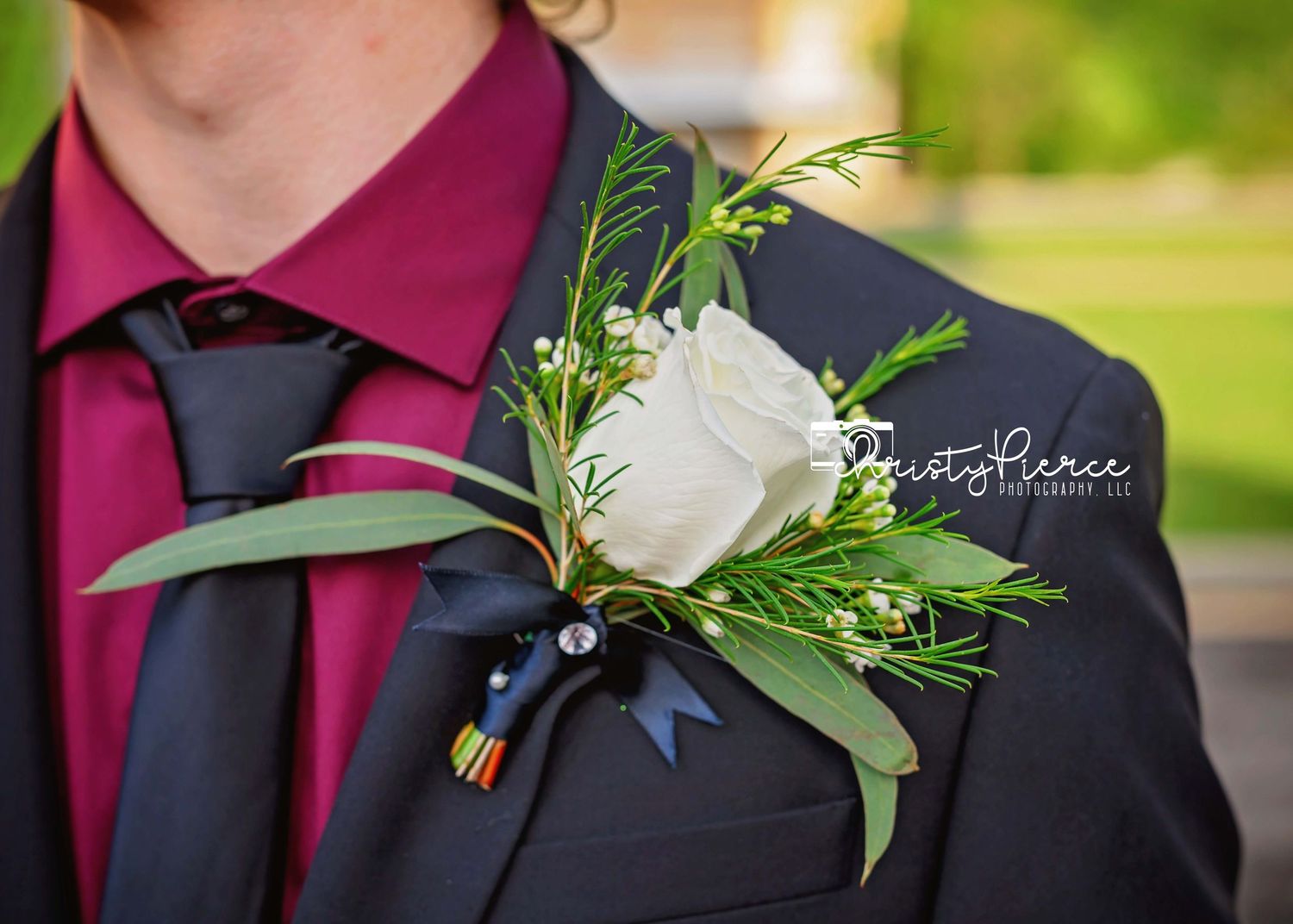 Wedding boutonniere made with white rose and greenery pinned to dark suit lapel against blurred outdoor background.