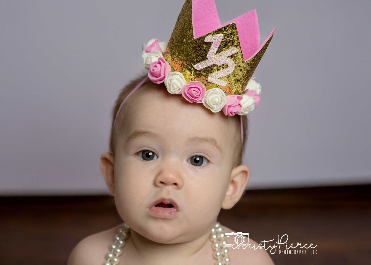 A small crown adorned with pink and gold decorations sits atop a head against a gray background.