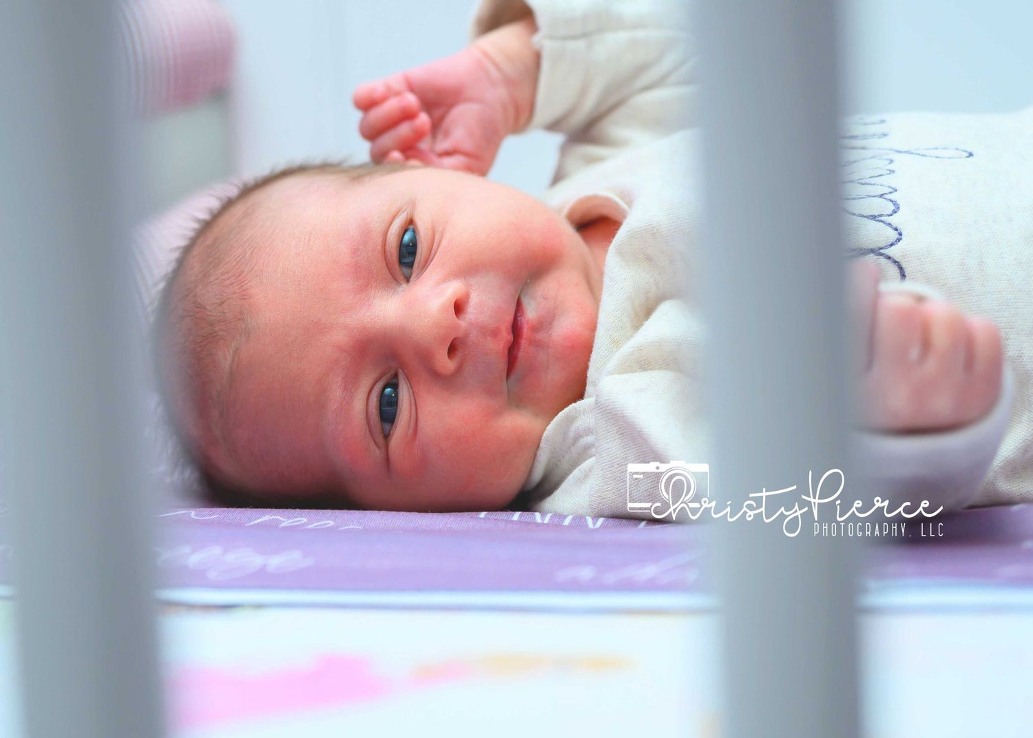 A newborn baby lies on pink bedding while gazing upward through white crib slats.