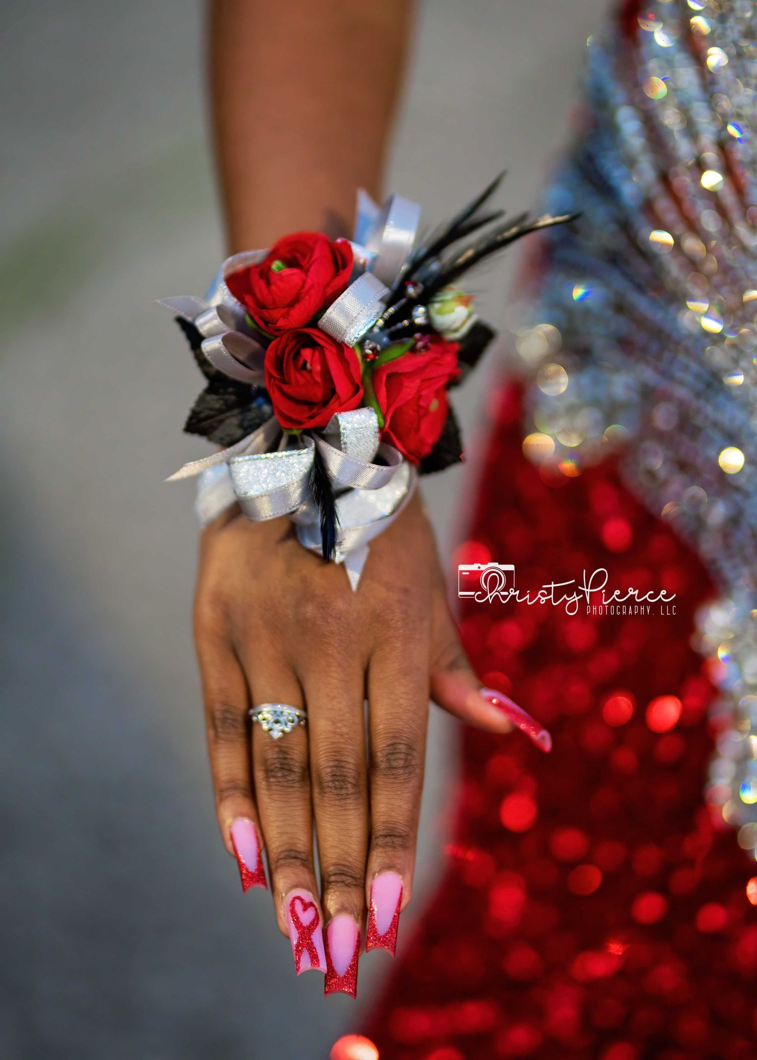 A hand adorned with pink nails displays a wrist corsage featuring vibrant red roses and silver accents against a sparkly backdrop.