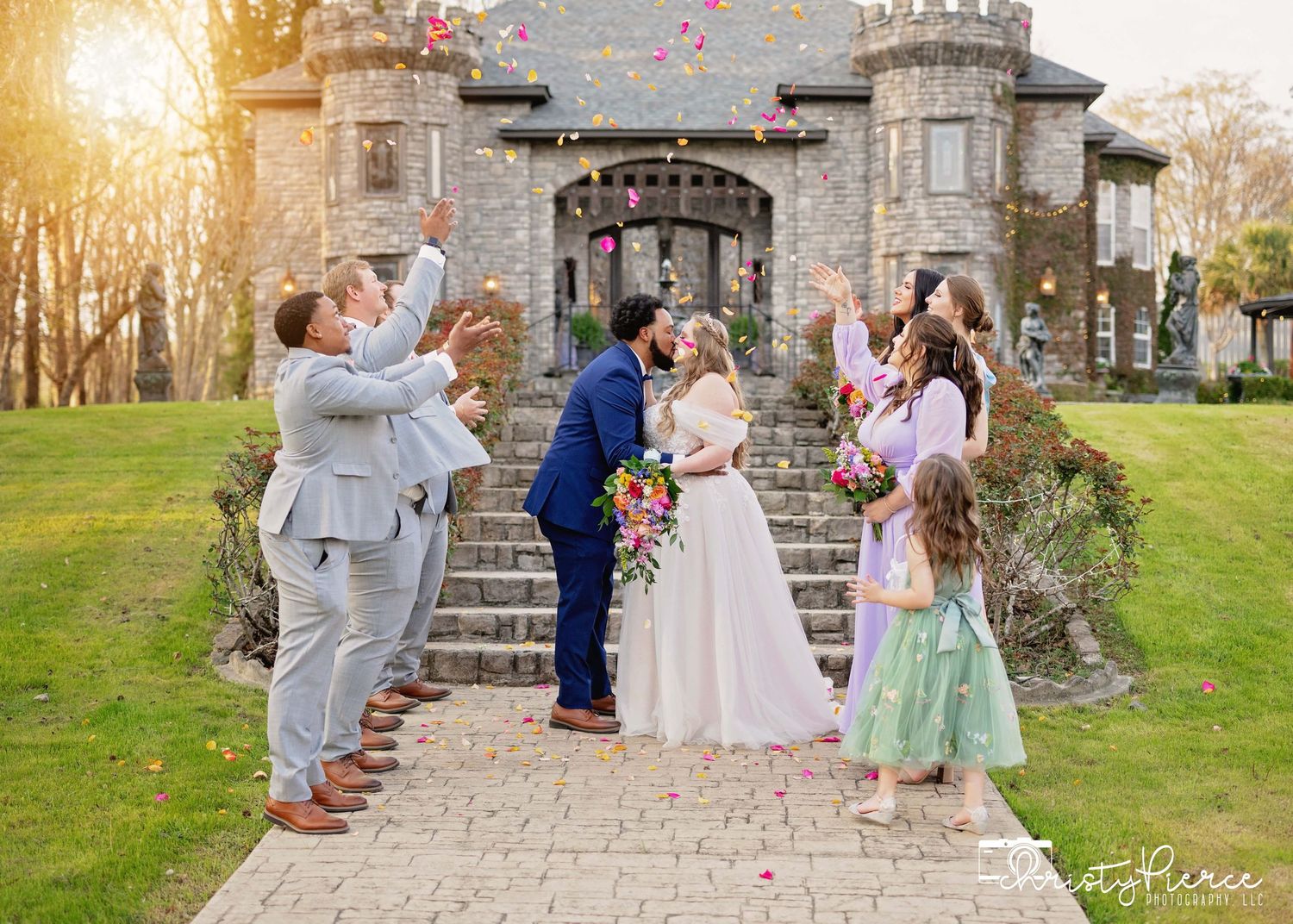 A romantic outdoor wedding ceremony with pink flower petals falling at a historic castle venue at sunset.