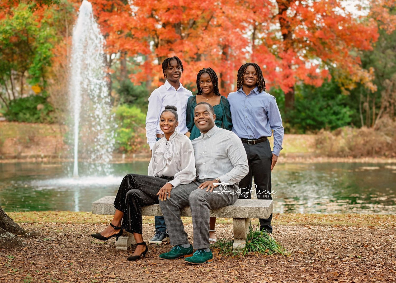 A family sits together outdoors during autumn with vibrant orange foliage and a water fountain in the background.