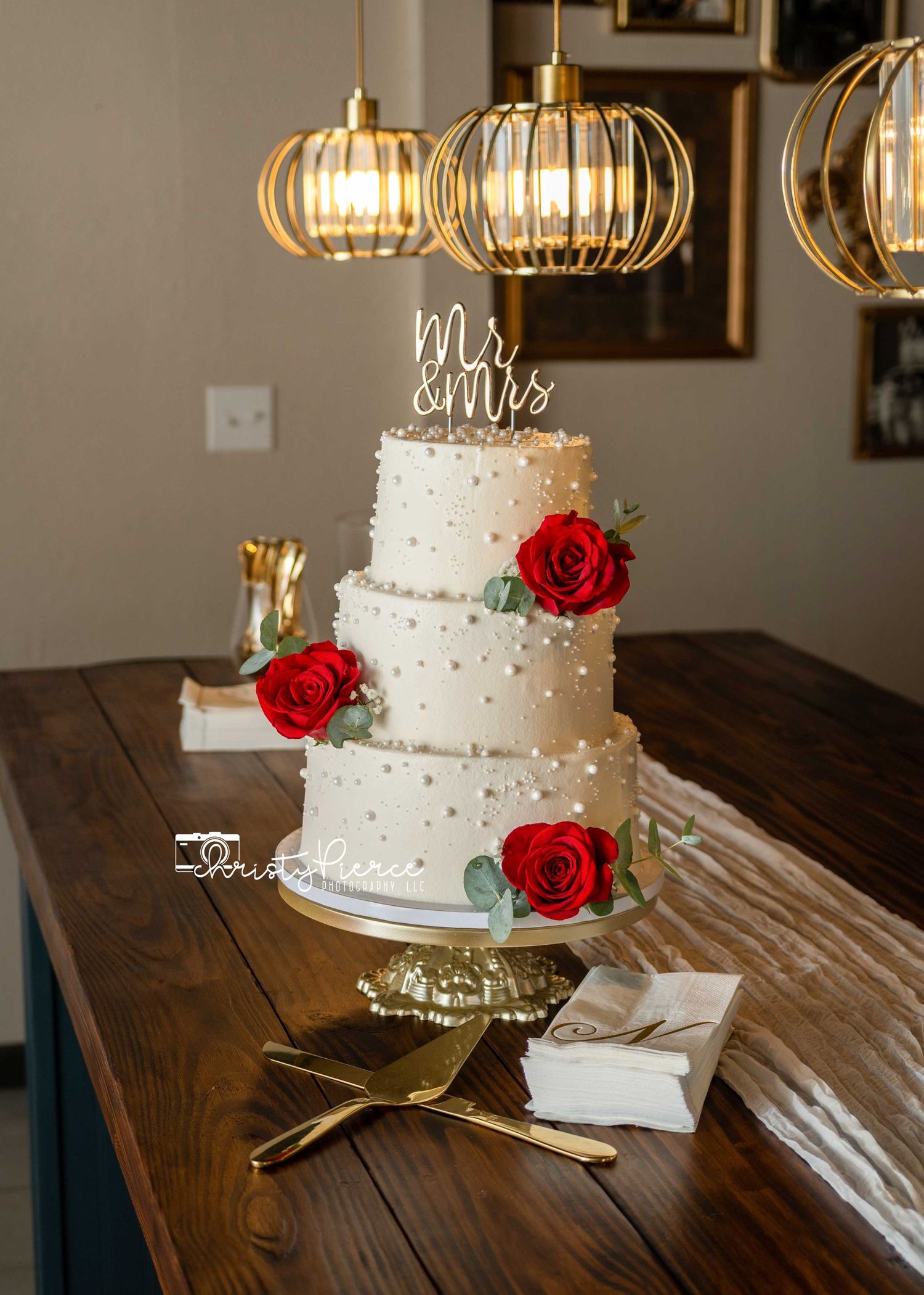 Three-tier white wedding cake with red roses and Mr & Mrs topper on gold stand under pendant lights.