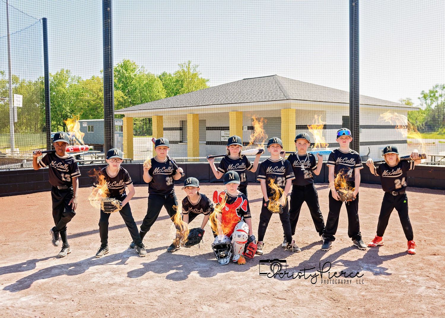 Youth baseball team poses in matching black uniforms on a softball field with dugout and buildings in background.