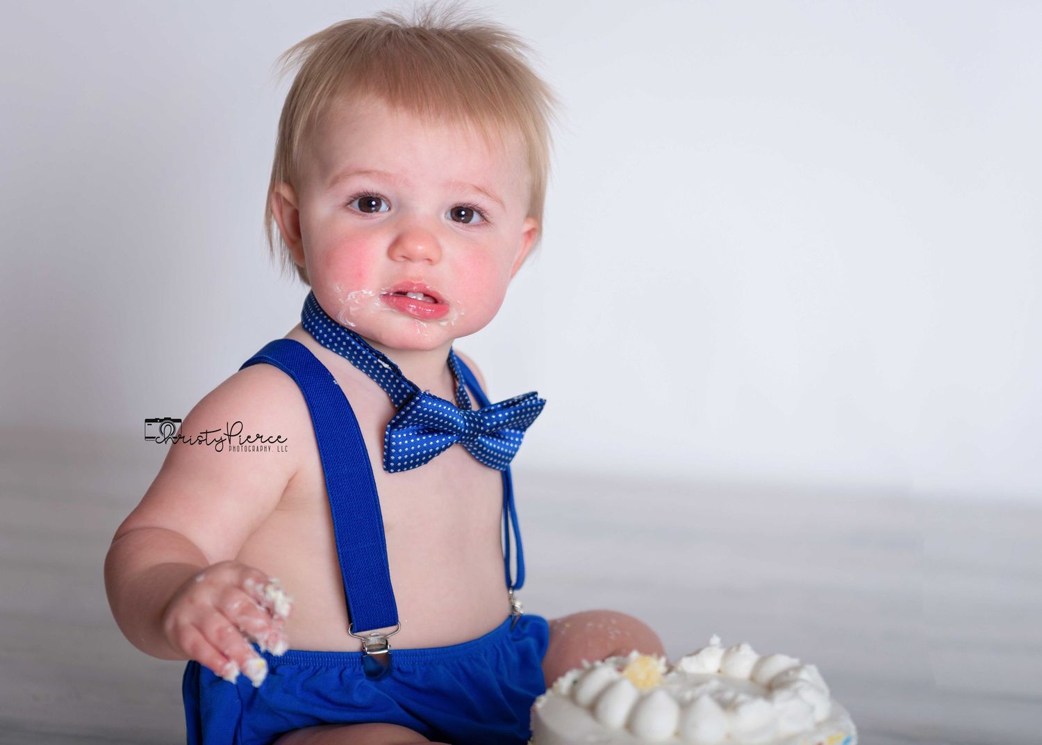 Baby wearing blue bow tie and suspenders sits beside white frosted cake for smash cake photo session.