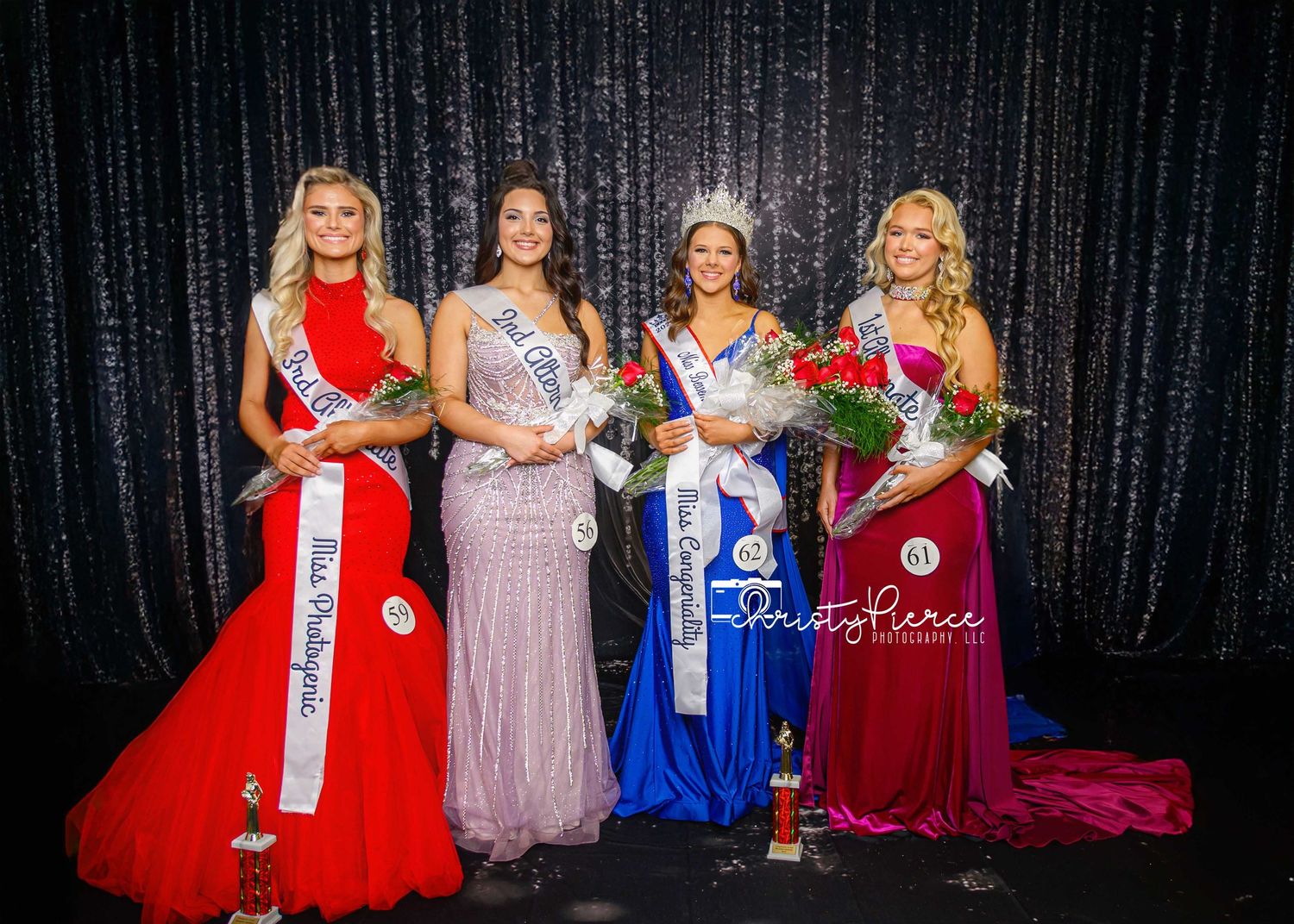 Pageant contestants in formal evening gowns pose together against a dark sparkly backdrop during a competition event.