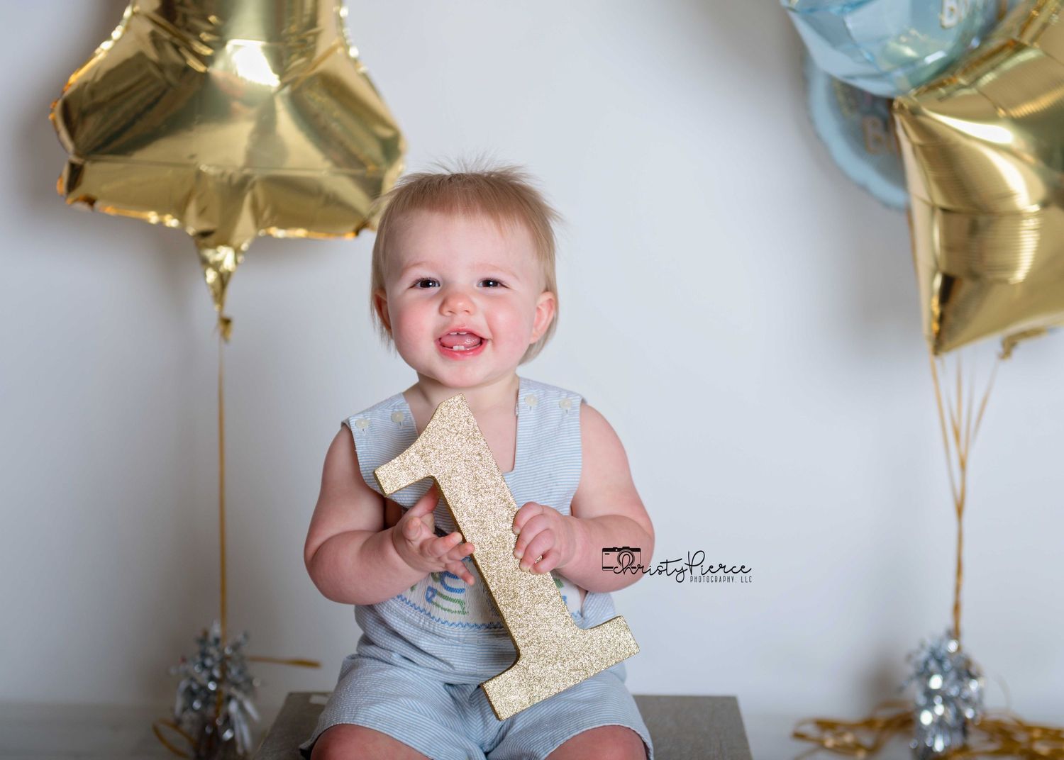 Baby in light blue outfit sits on floor with gold star balloons during first birthday photoshoot celebration.