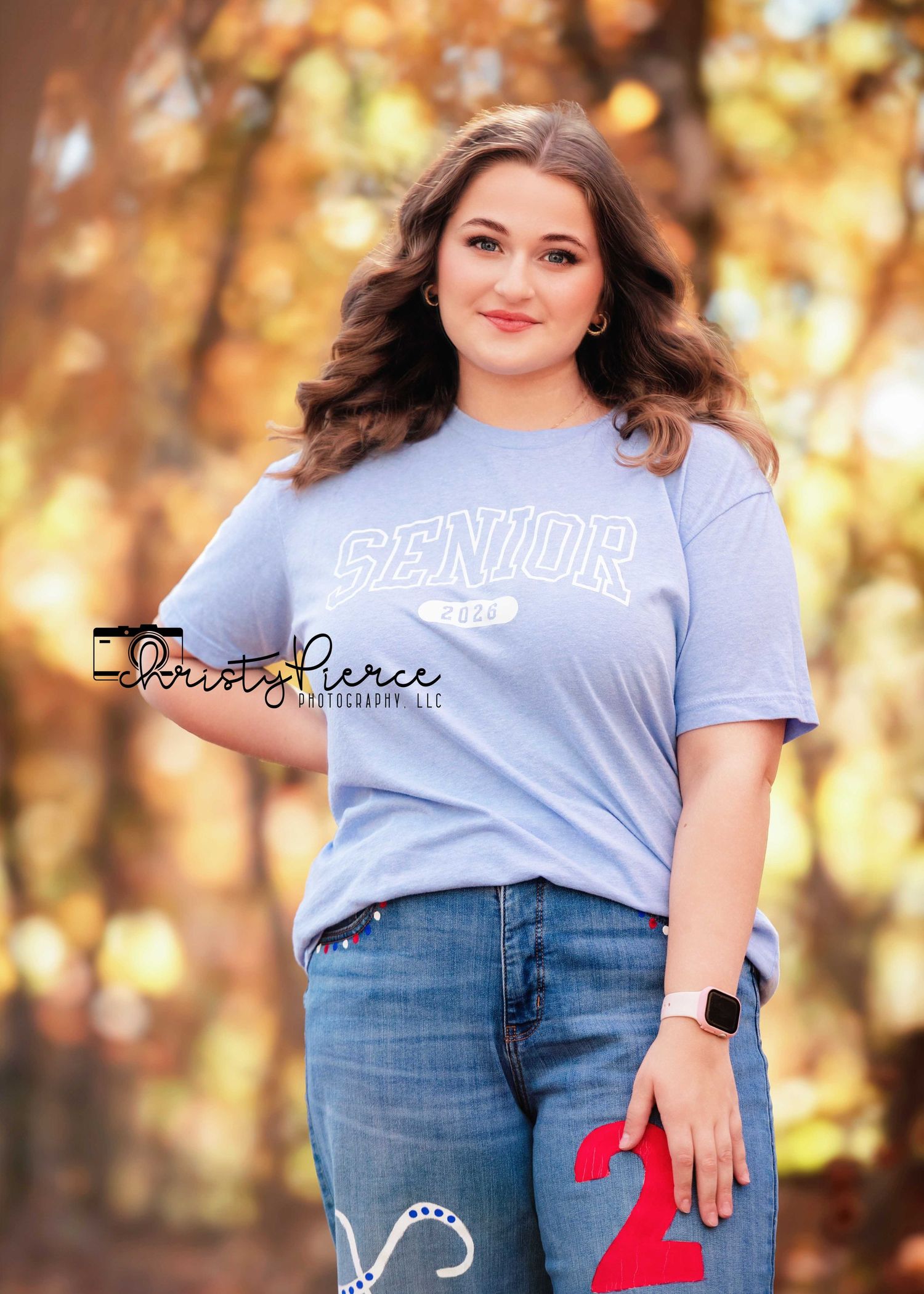 A young woman in a blue Senior 2025 t-shirt and jeans poses outdoors among autumn foliage for senior portraits.