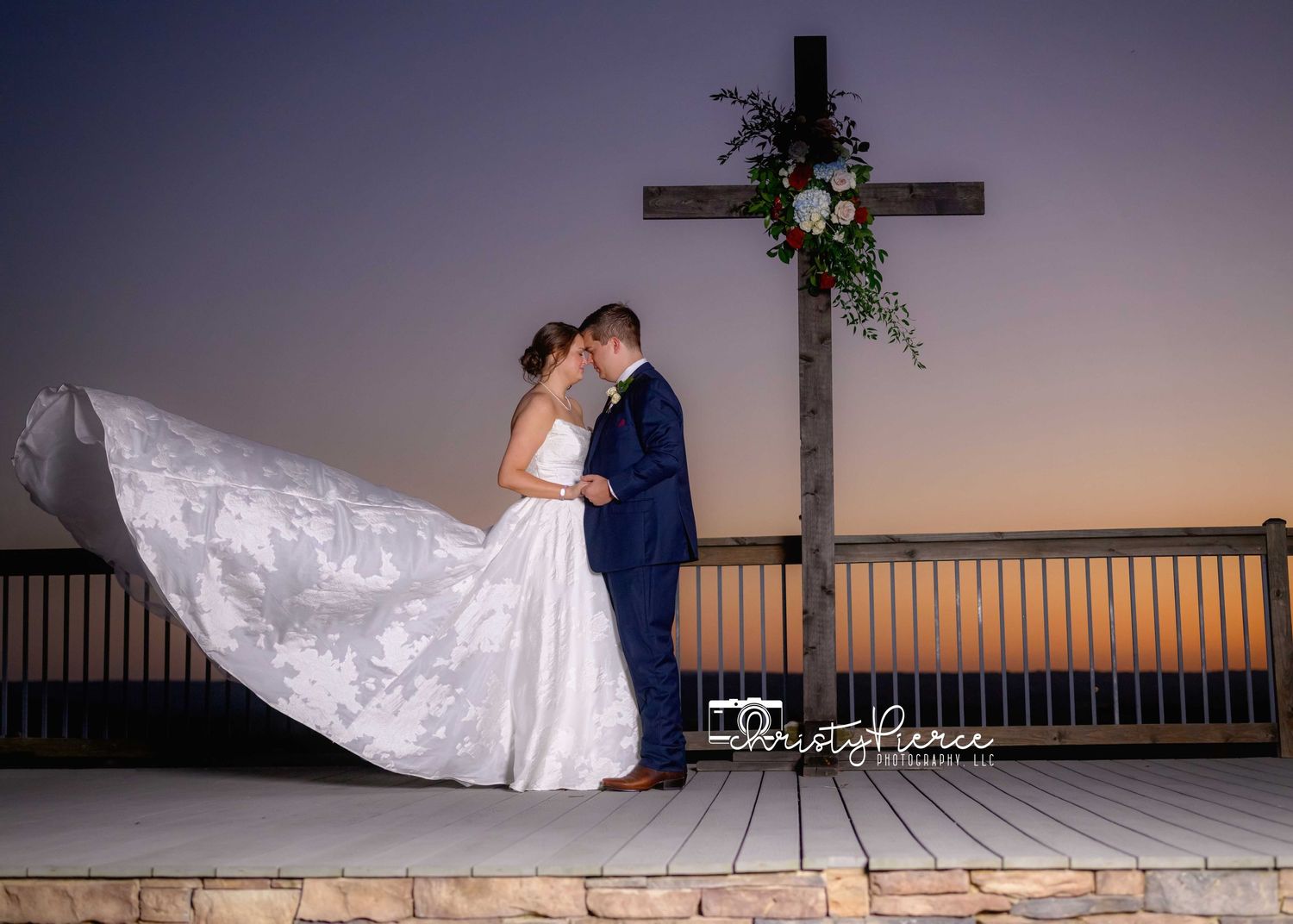 Bride and groom share a romantic kiss at sunset beside a wooden cross decorated with flowers on an outdoor deck.