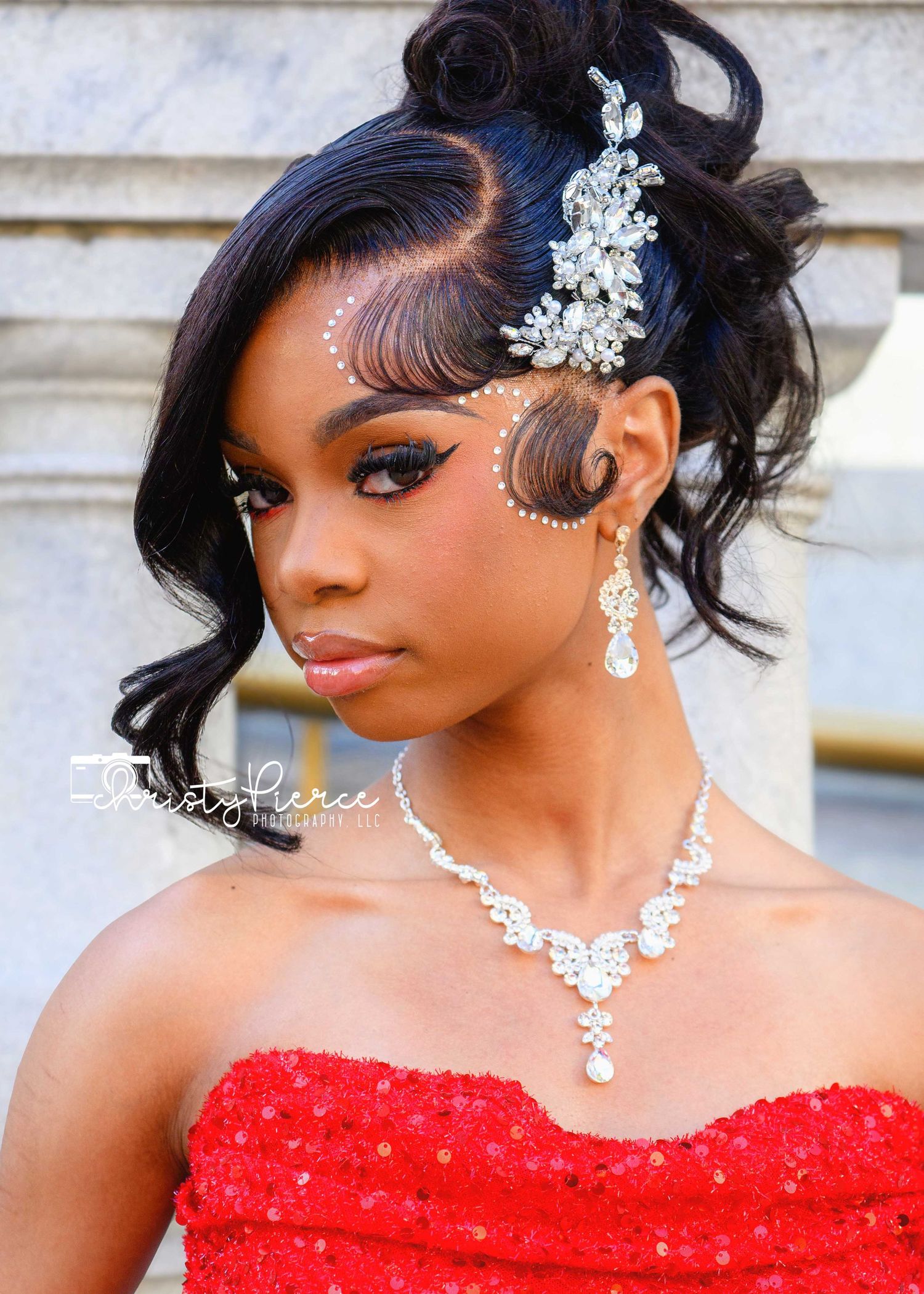 Young woman in red sequin dress with crystal hair piece, rhinestone jewelry, and elegant updo hairstyle for formal event.