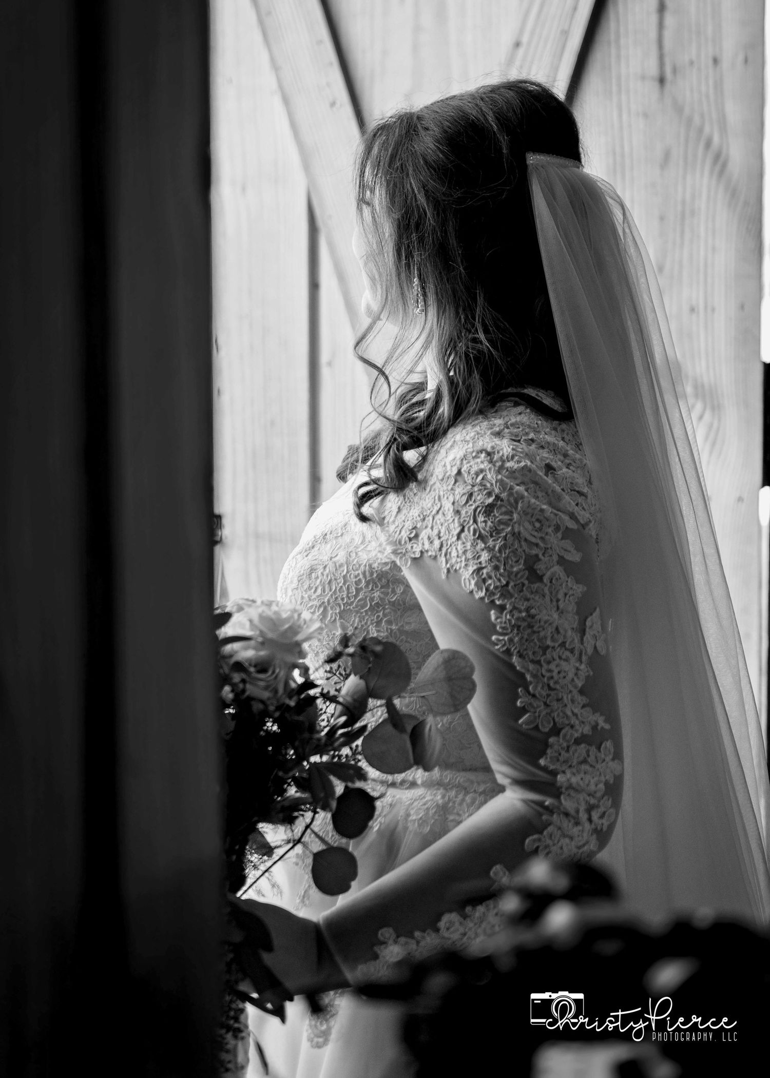 Black and white photo of a bride in a lace wedding dress with long veil holding a bouquet by a window.