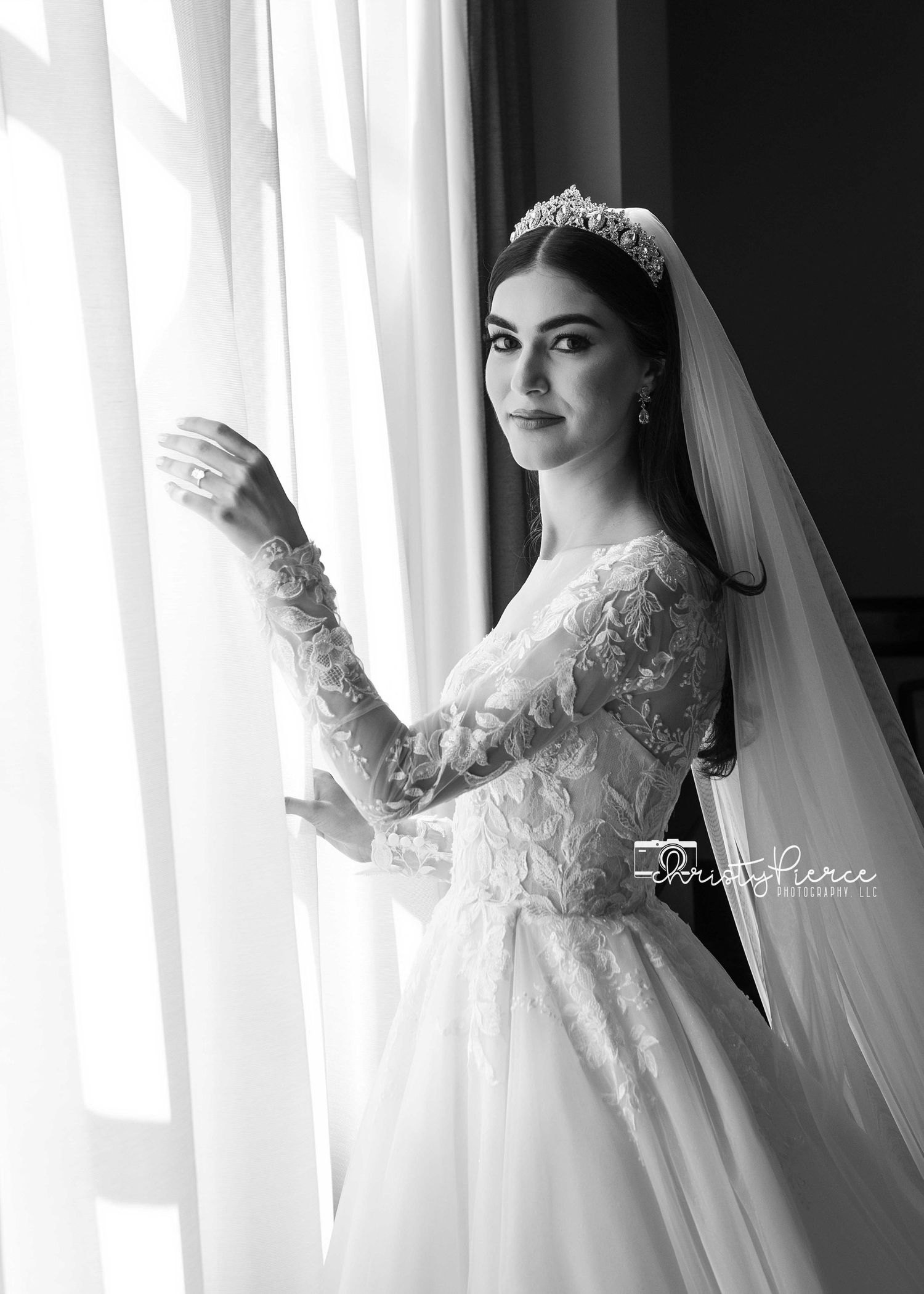 A bride in an elegant long-sleeved lace wedding gown and veil poses gracefully against white curtains in a black and white photo.