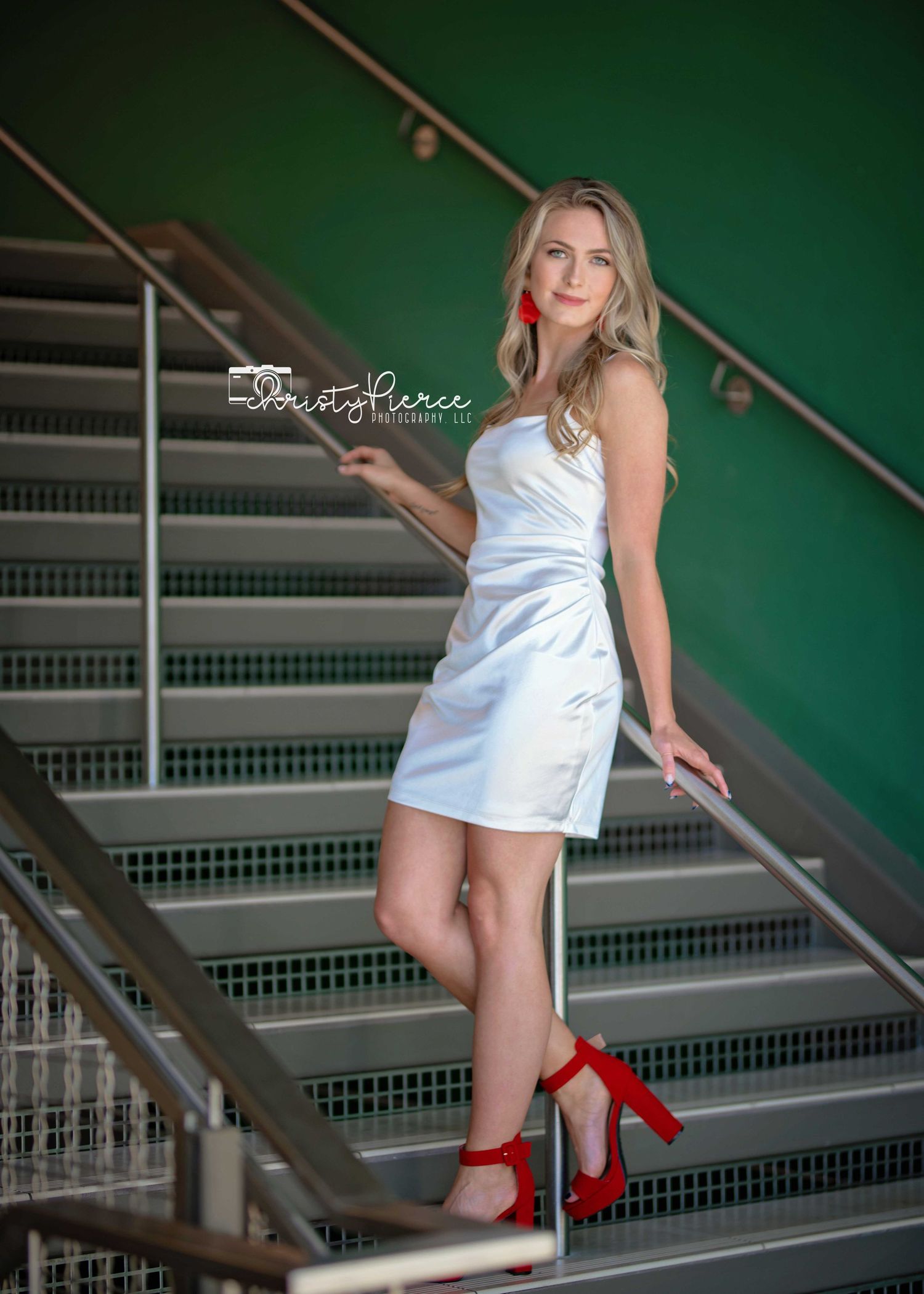 Young woman in white dress and red heels poses gracefully on modern stairs with green wall backdrop.