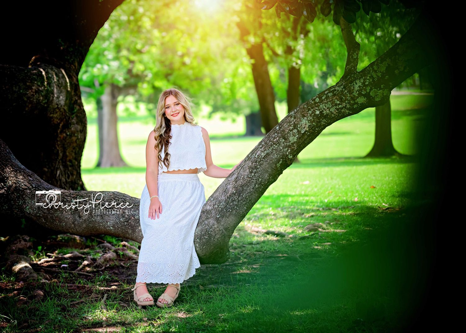 Young woman in white maxi dress leaning against a large tree in a lush green park, bathed in warm sunlight.