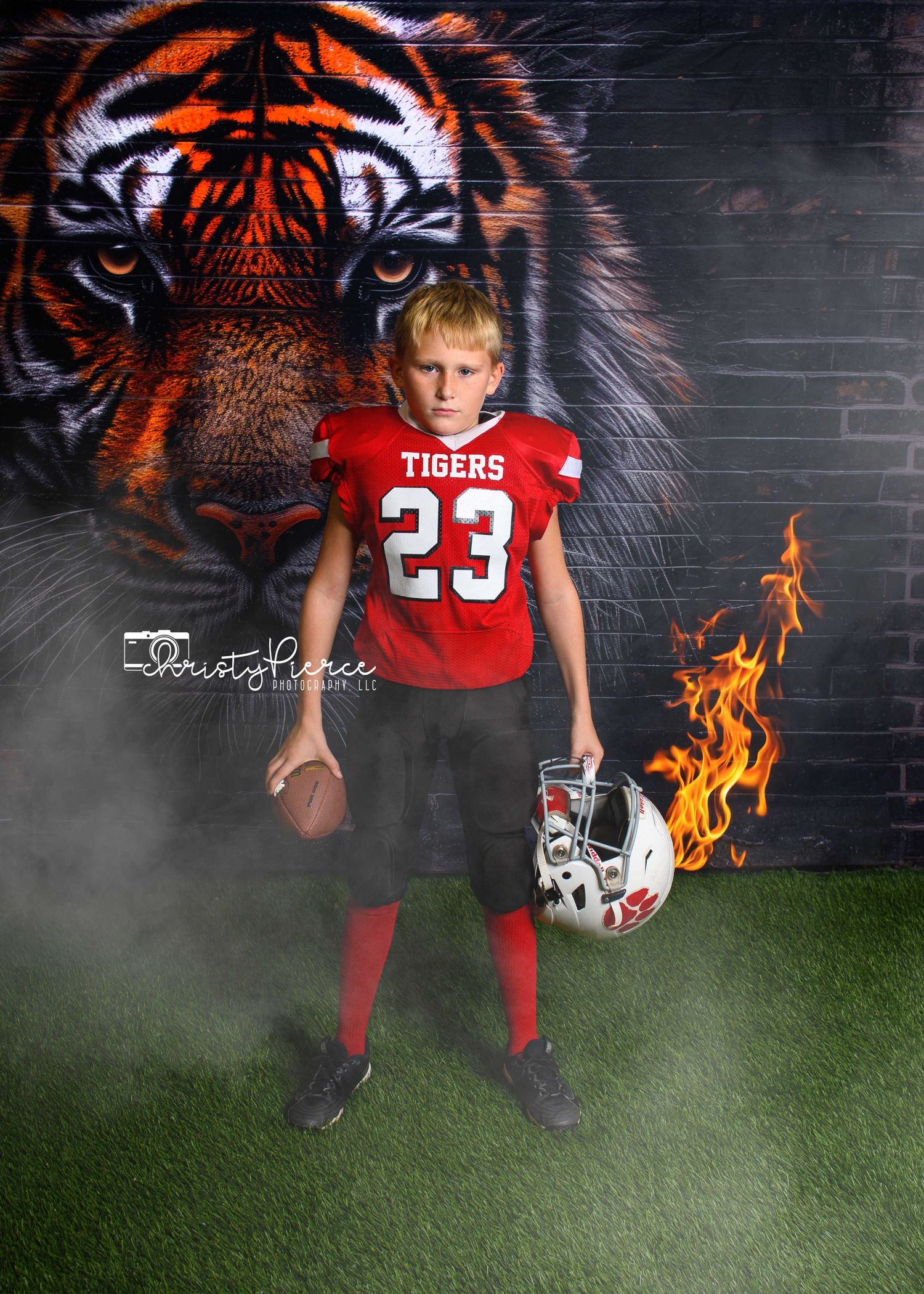 Young football player in red Tigers #23 jersey poses with helmet and ball in front of tiger mural.