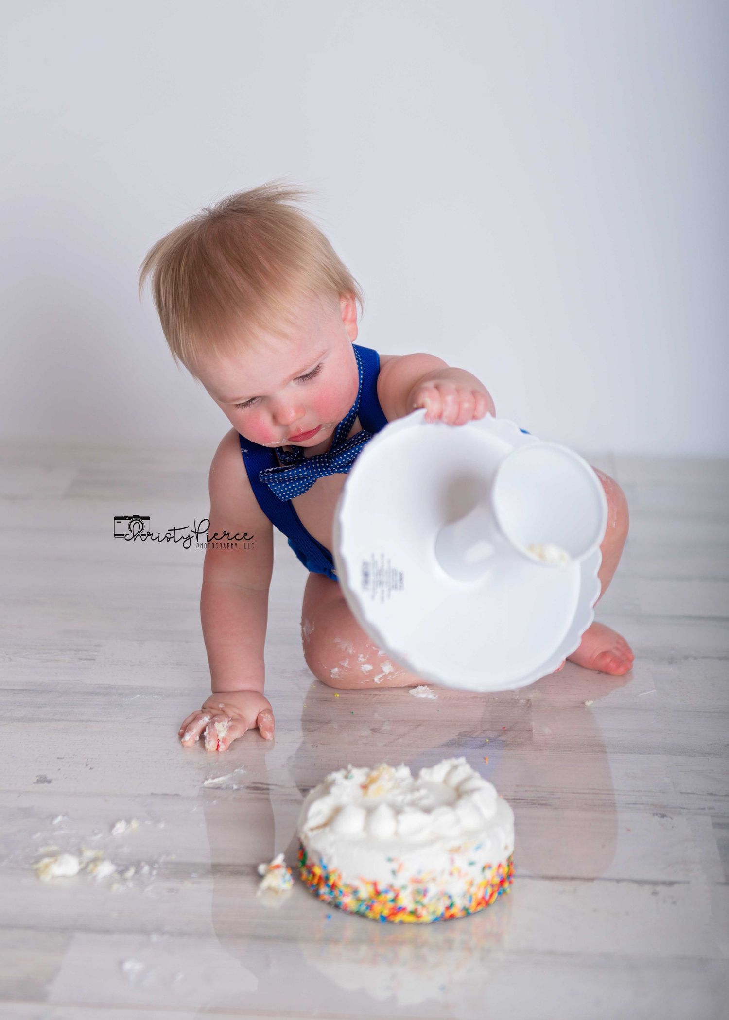 A baby in a blue shirt crawls and plays with a white plate and scattered food on a light wooden floor.