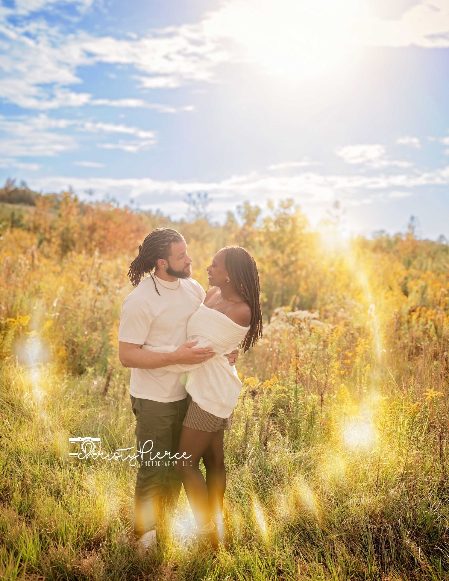 Couple embraces in a sunlit meadow with tall grass and golden light during sunset.