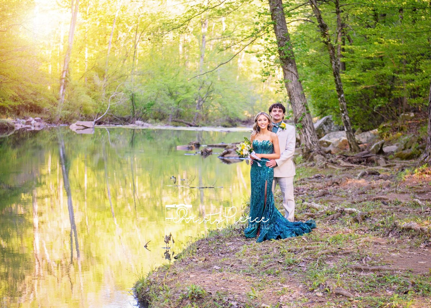 A couple in elegant formal attire stands by a tranquil pond in a sunlit forest setting during golden hour.