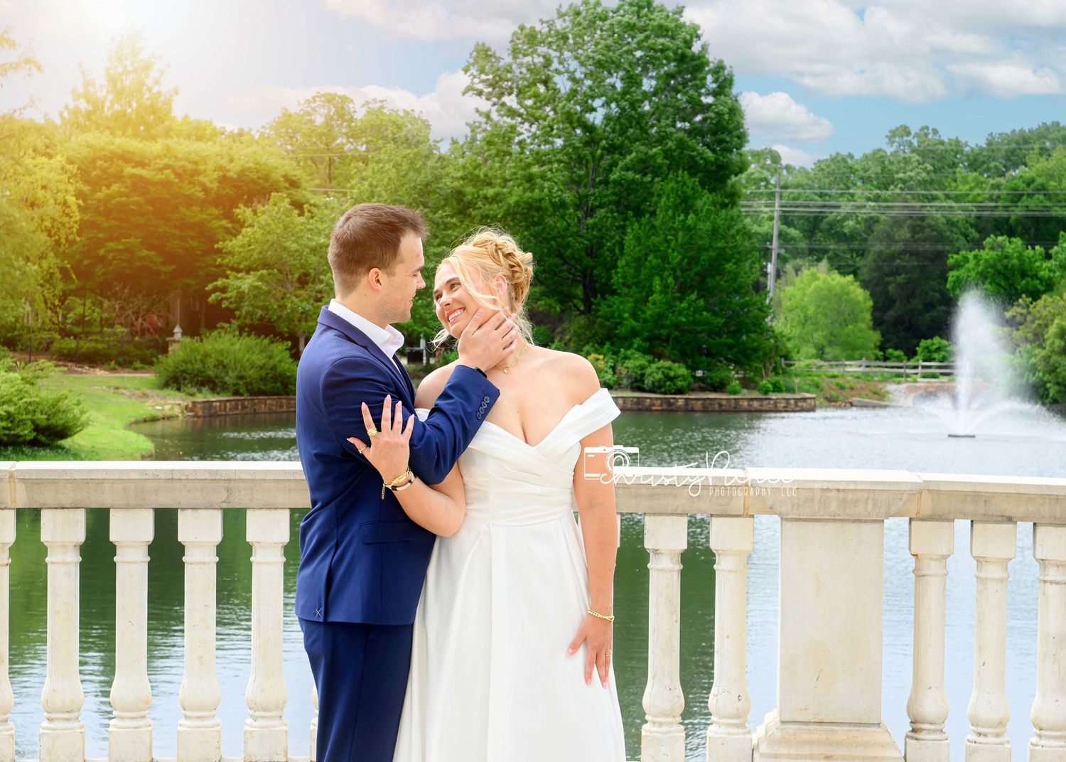 Bride and groom embrace on stone balcony overlooking fountain and pond at outdoor garden wedding.