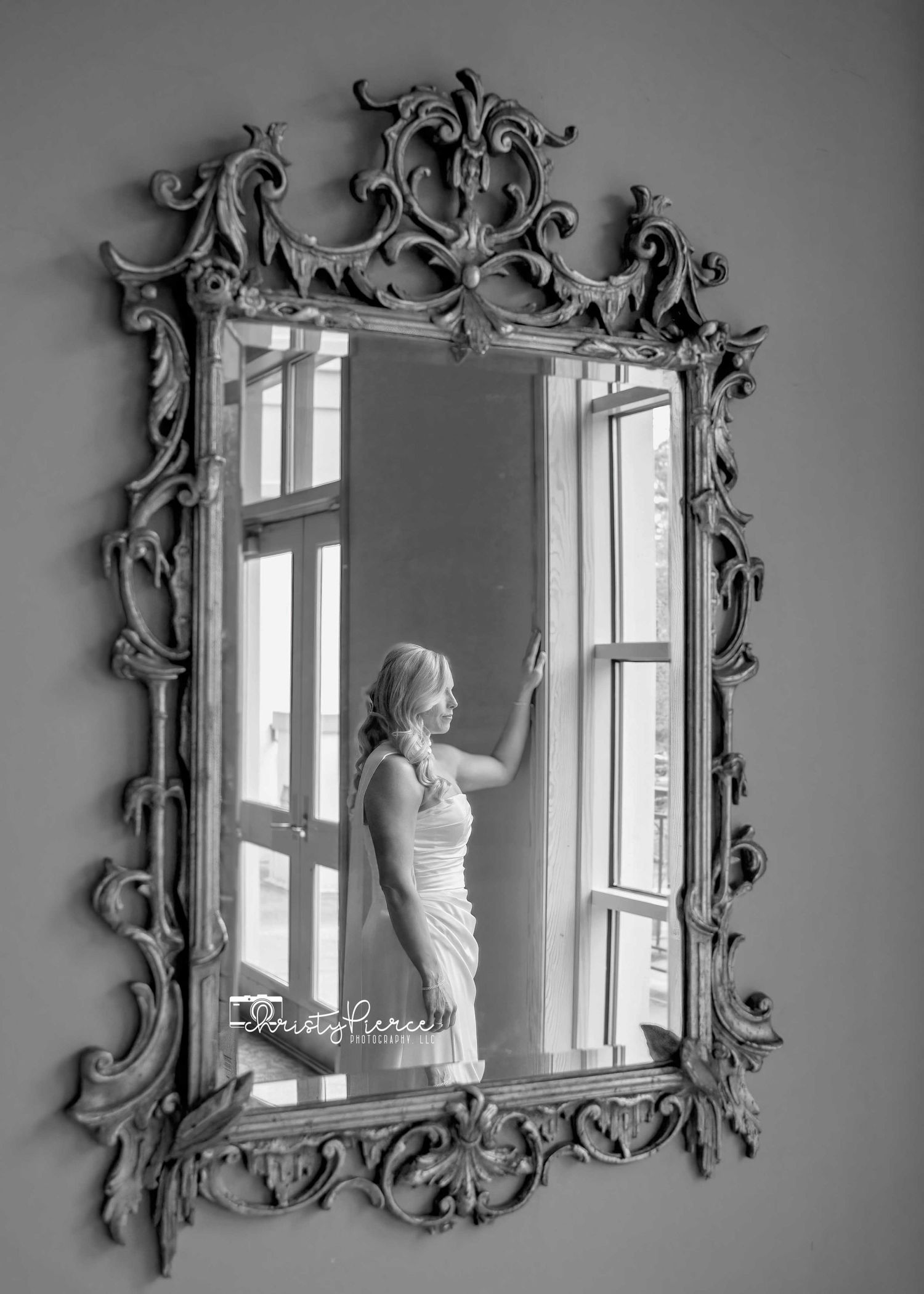Black and white photo of a bride in a white dress reflected in an ornate vintage mirror near a bright window.