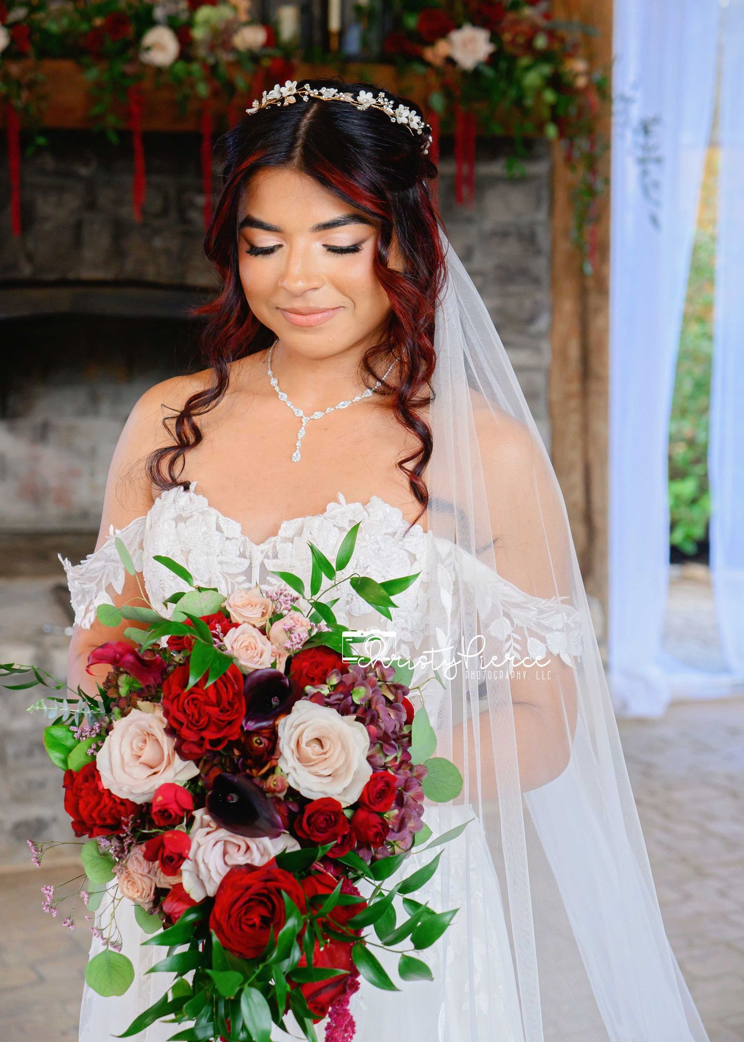 Bride in white lace gown holds cascading red and blush rose bouquet, wearing veil and pearl necklace.