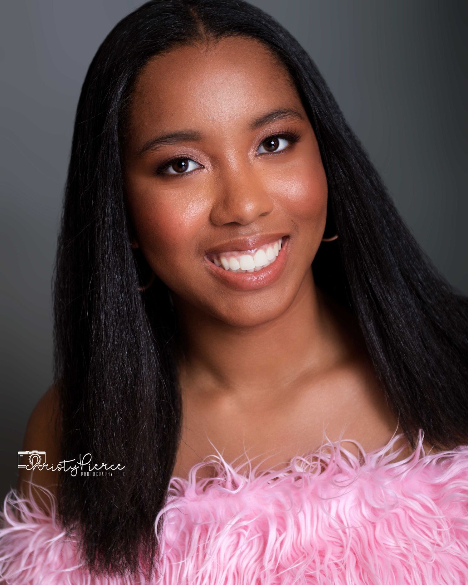 Portrait against dark background wearing a pink feathered top and long straight black hair with a bright smile.
